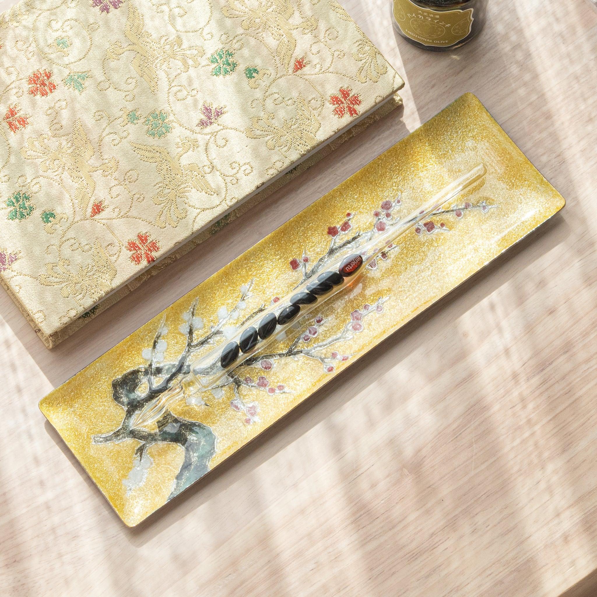 Angled view of gold tray with plum blossom design and glass pen, alongside a decorative notebook on a wooden surface.