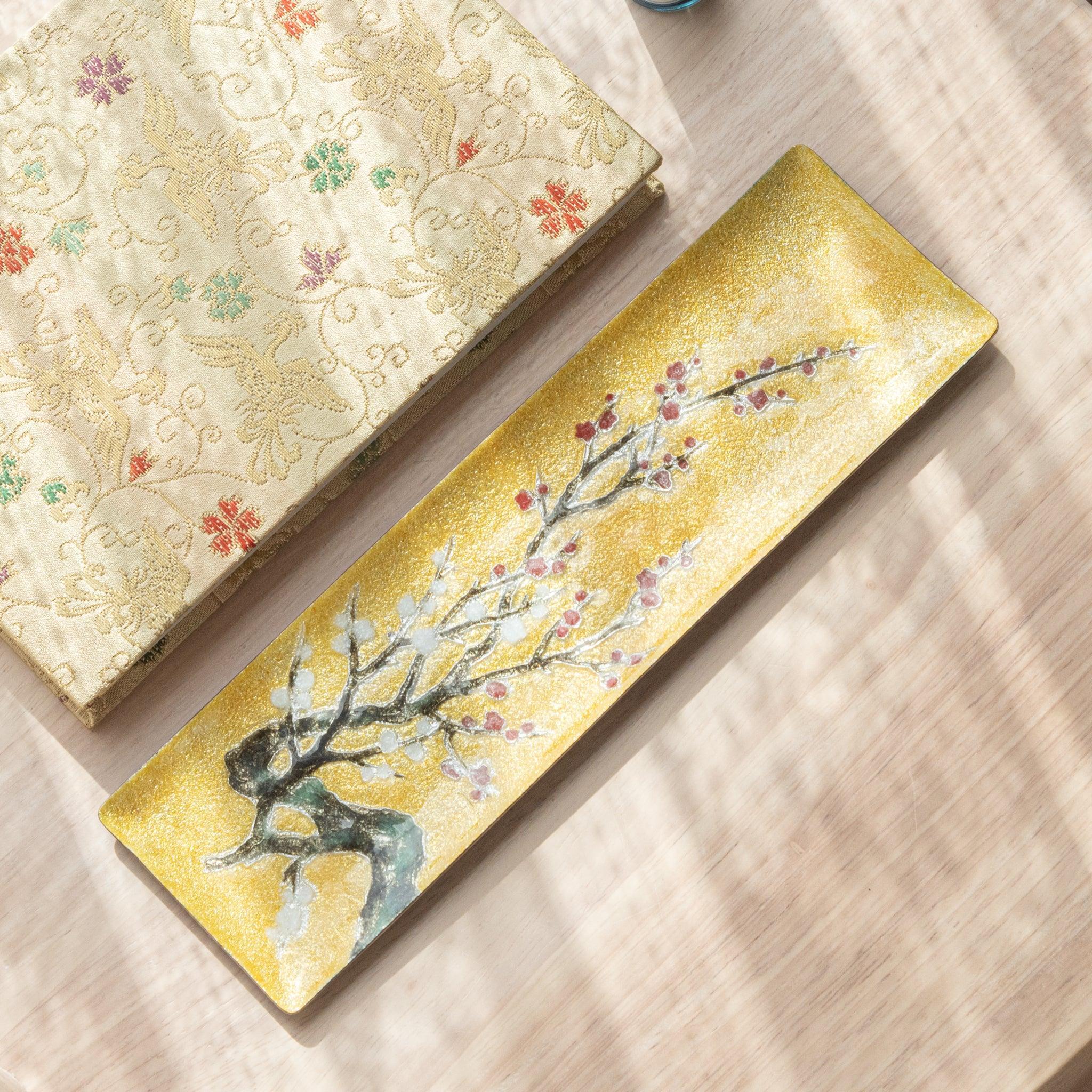 Close-up of gold tray featuring intricate plum blossom design with a glass pen resting on it, showcasing fine texture and detail on a wooden surface.