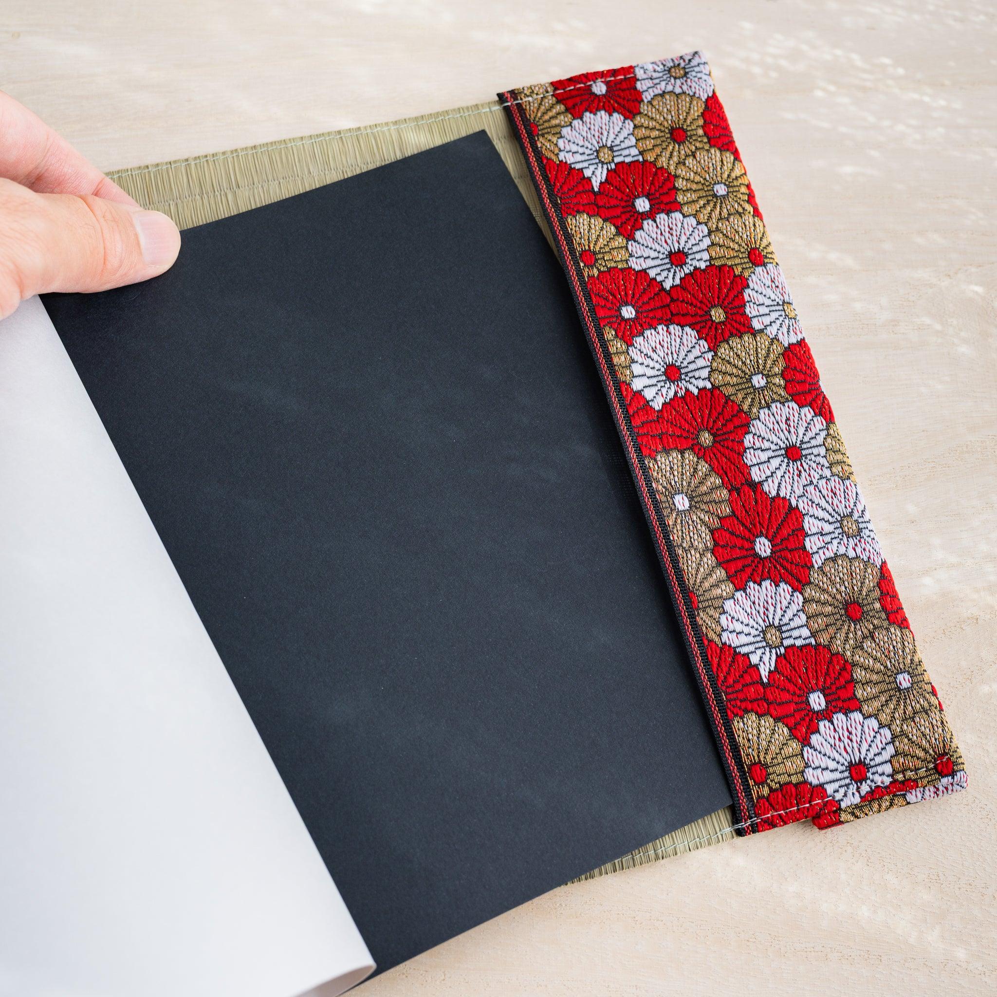 Hand lifting page inside tatami book cover, black notebook cover visible beside red and gold chrysanthemum fabric band
