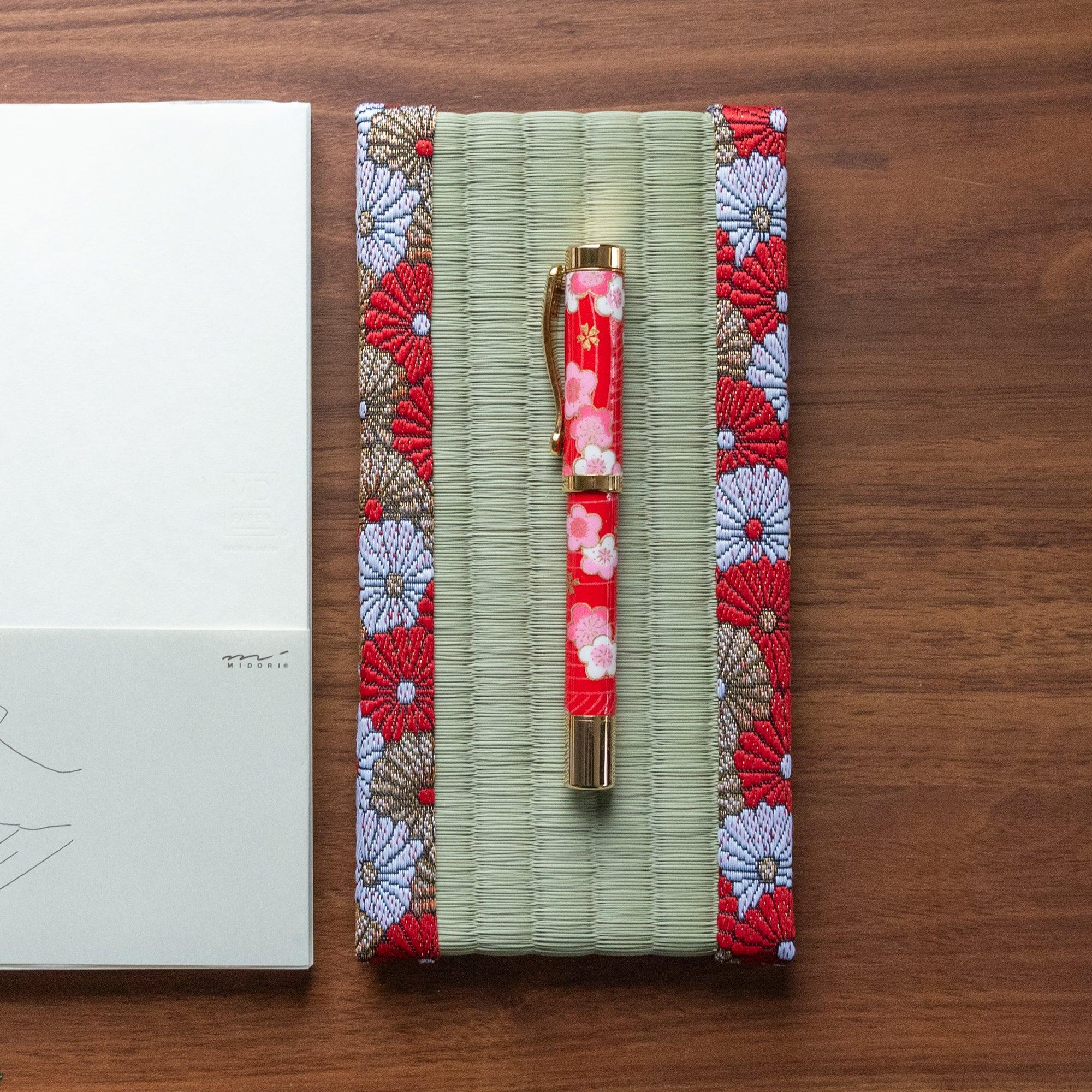 Red and gold floral Japanese tatami pen tray holding a decorative red pen with floral patterns, placed on a wooden surface next to a notebook.  