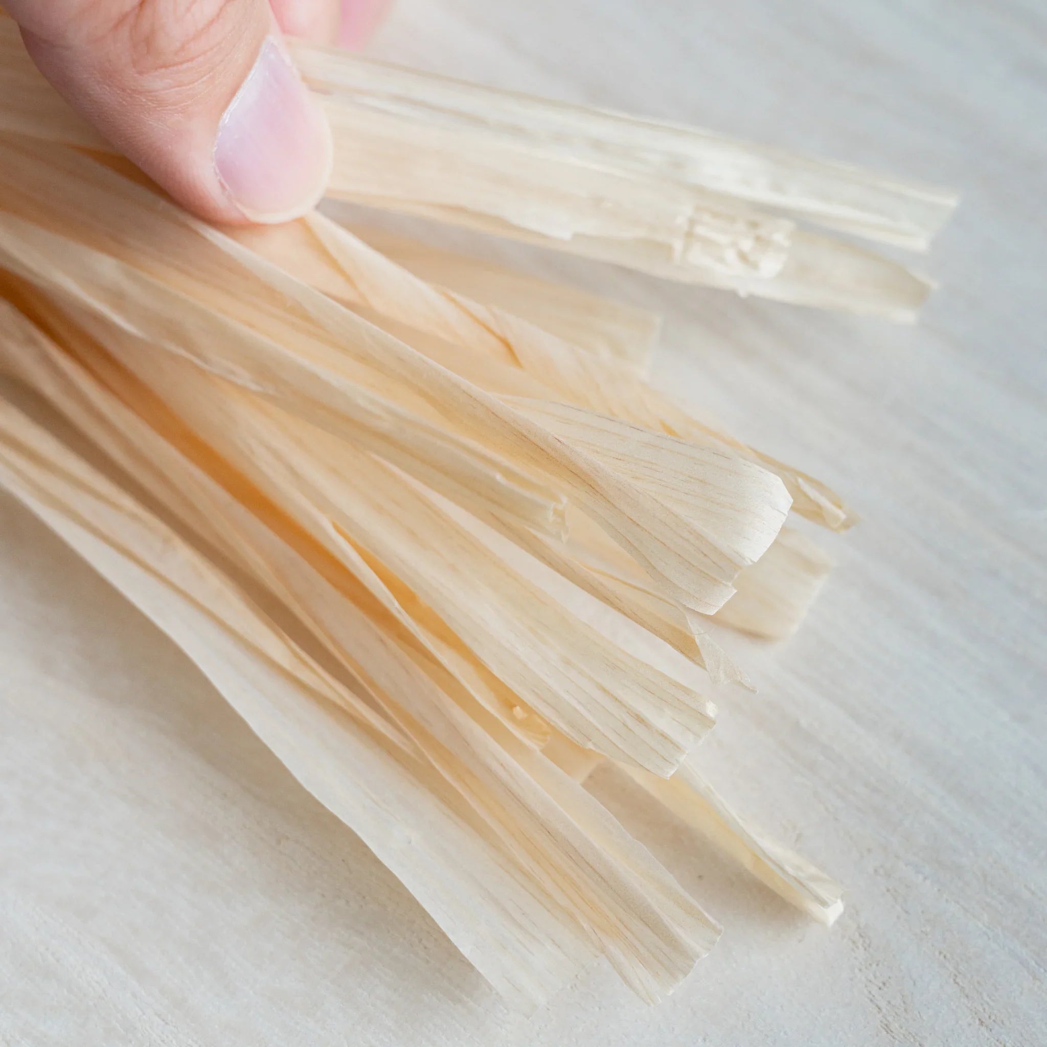 Hand holding thin ribbon-like Hinoki wood strings, pale wood surface below