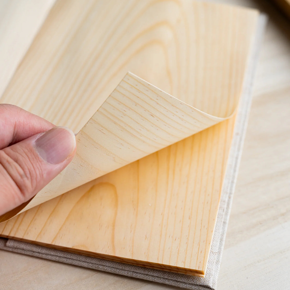 Close-up of a wood sheet page corner being lifted by hand, grain lines running along the sheet