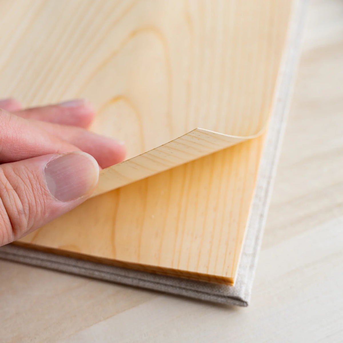 Thumb peeling back the corner of a thin wood sheet page, showing its flexibility and grain