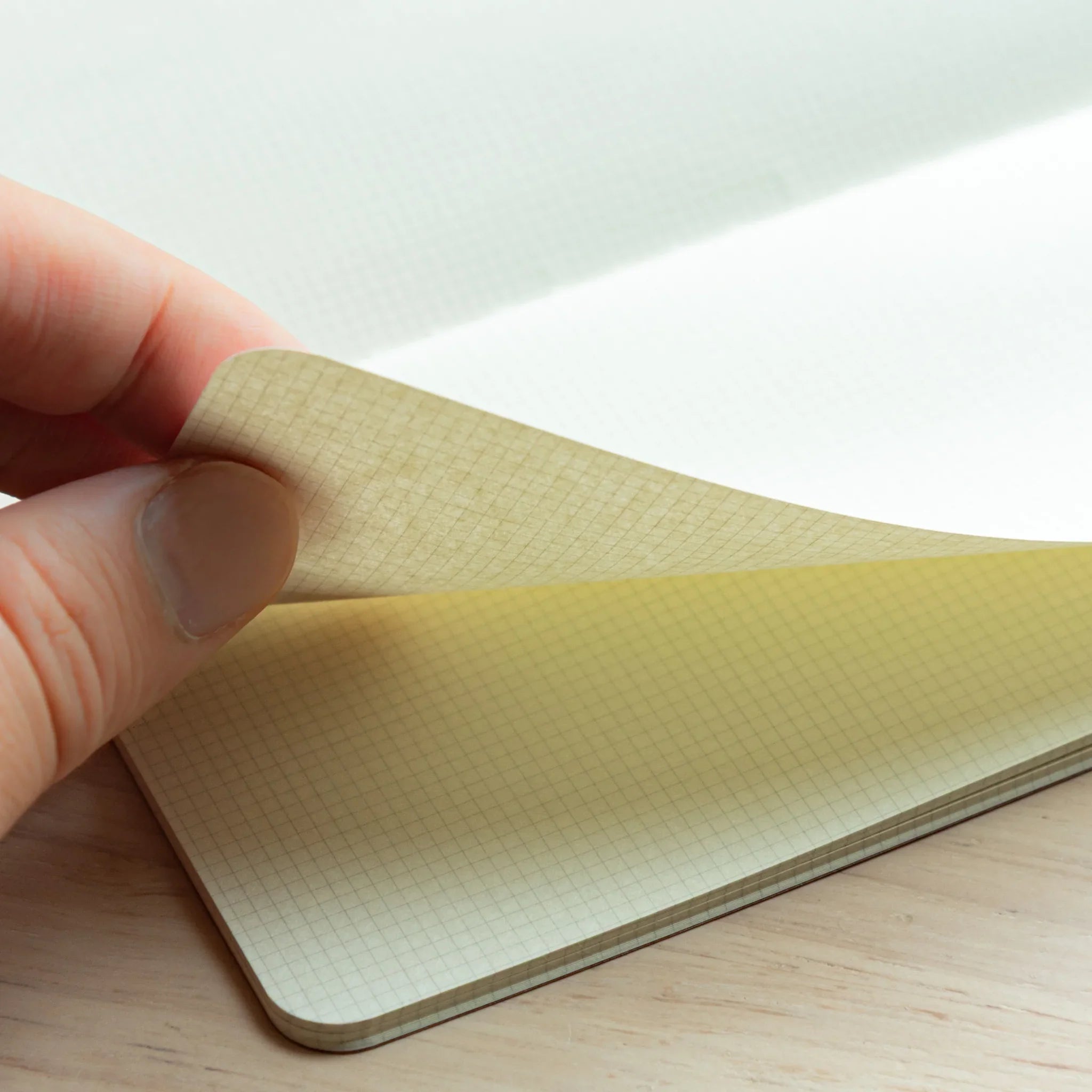 Hand lifting a page of the RO-BIKI Basic Series Japanese Notebook with a 2 mm grid, showing thin cream paper and square grid lines.