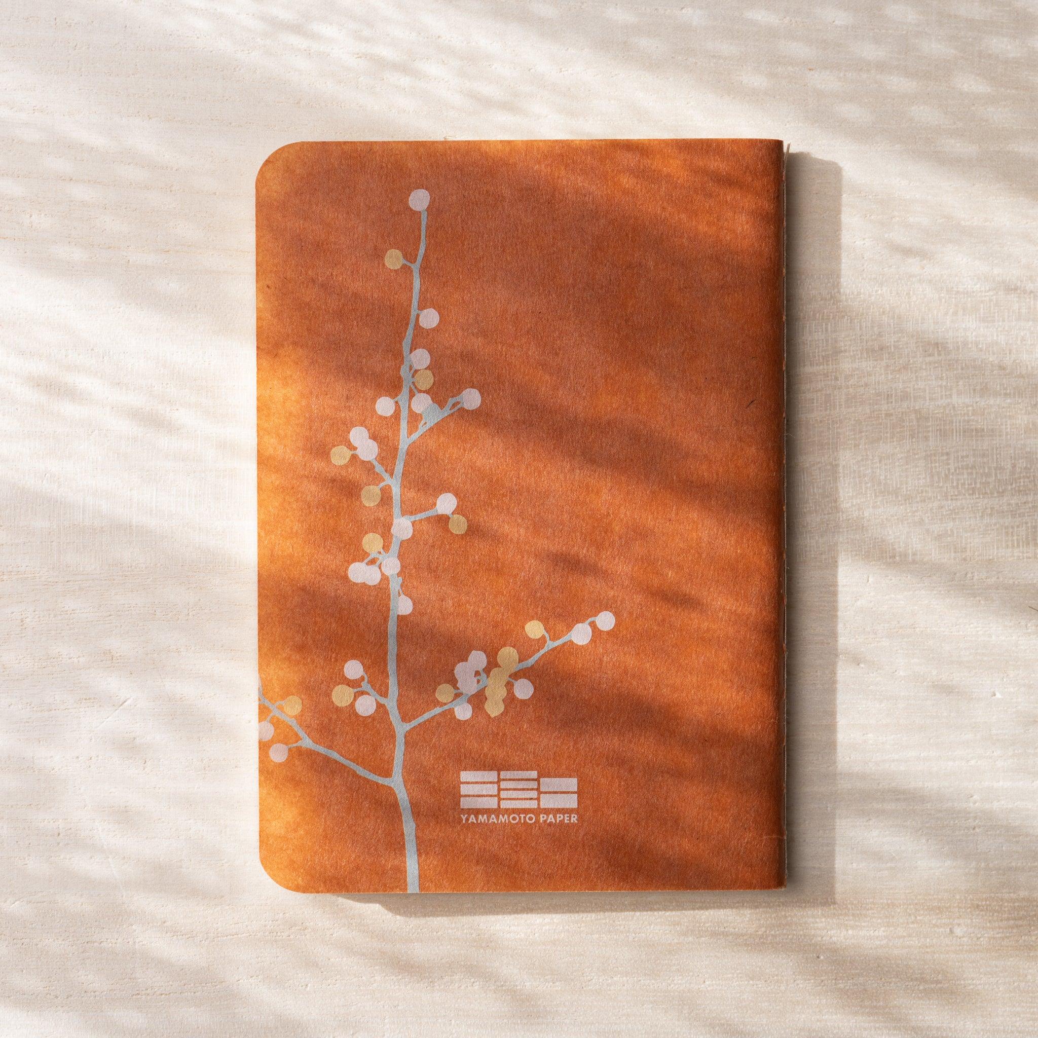 Close-up of RO-BIKI NOTE waxed cover showing branch and botanical flower pattern, terracotta tone visible