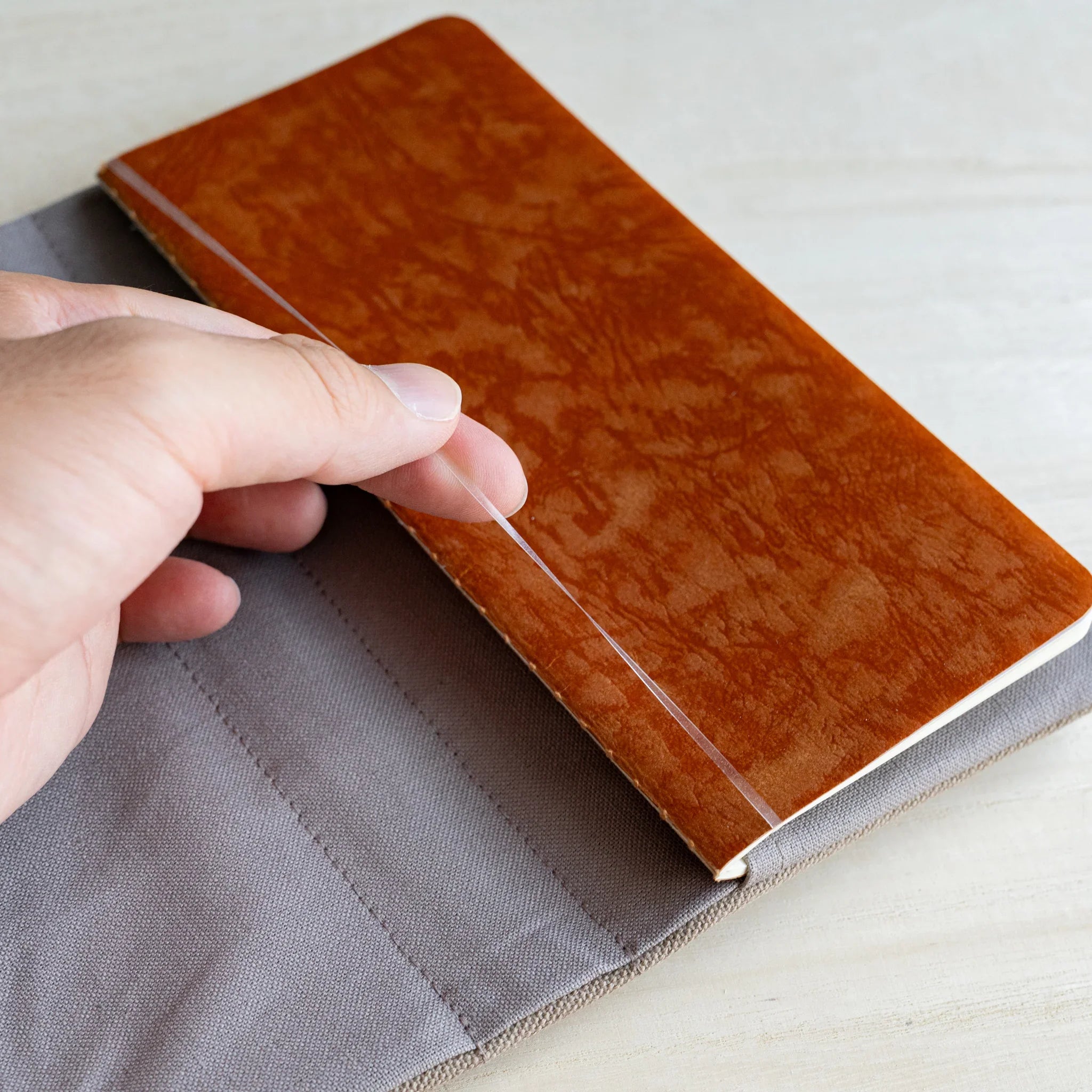 Hand sliding a tan waxed RO-BIKI notebook into the grey inner flap of the clay beige canvas cover