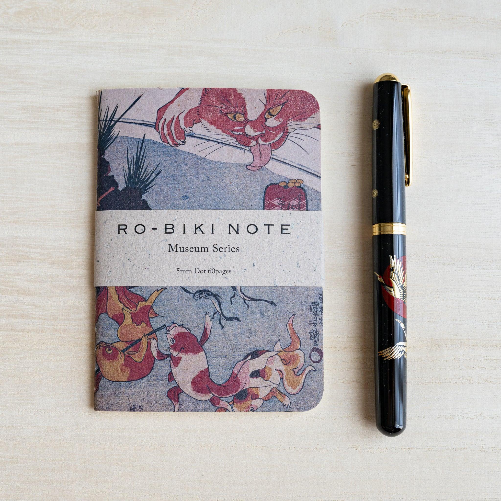 RO-BIKI Kingyo Goldfish notebook beside a black lacquer fountain pen with a gold crane design on pale wood