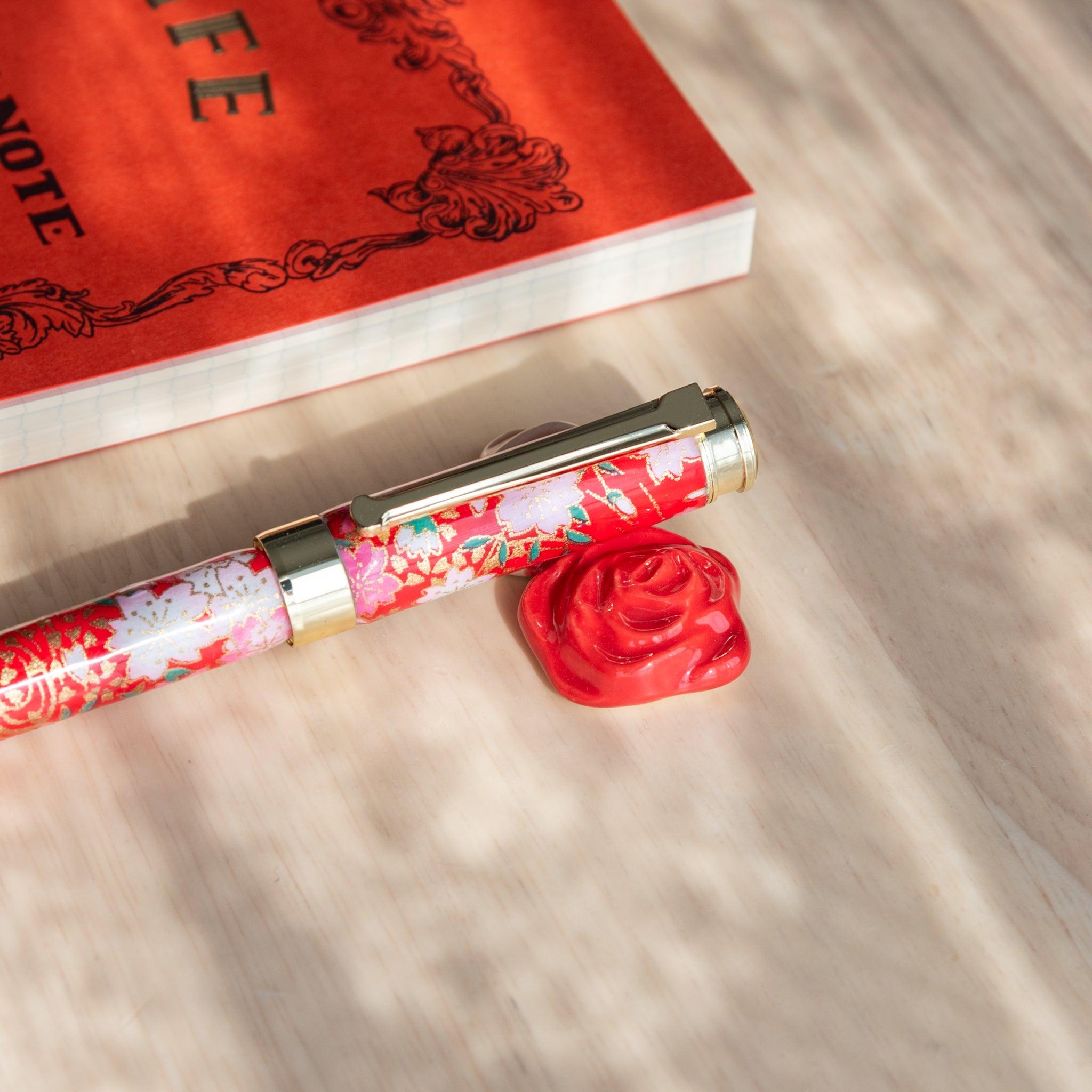 Floral Japanese fountain pen resting on a red and white rose-shaped ceramic pen rest, part of Japanese stationery, beside an orange notebook.