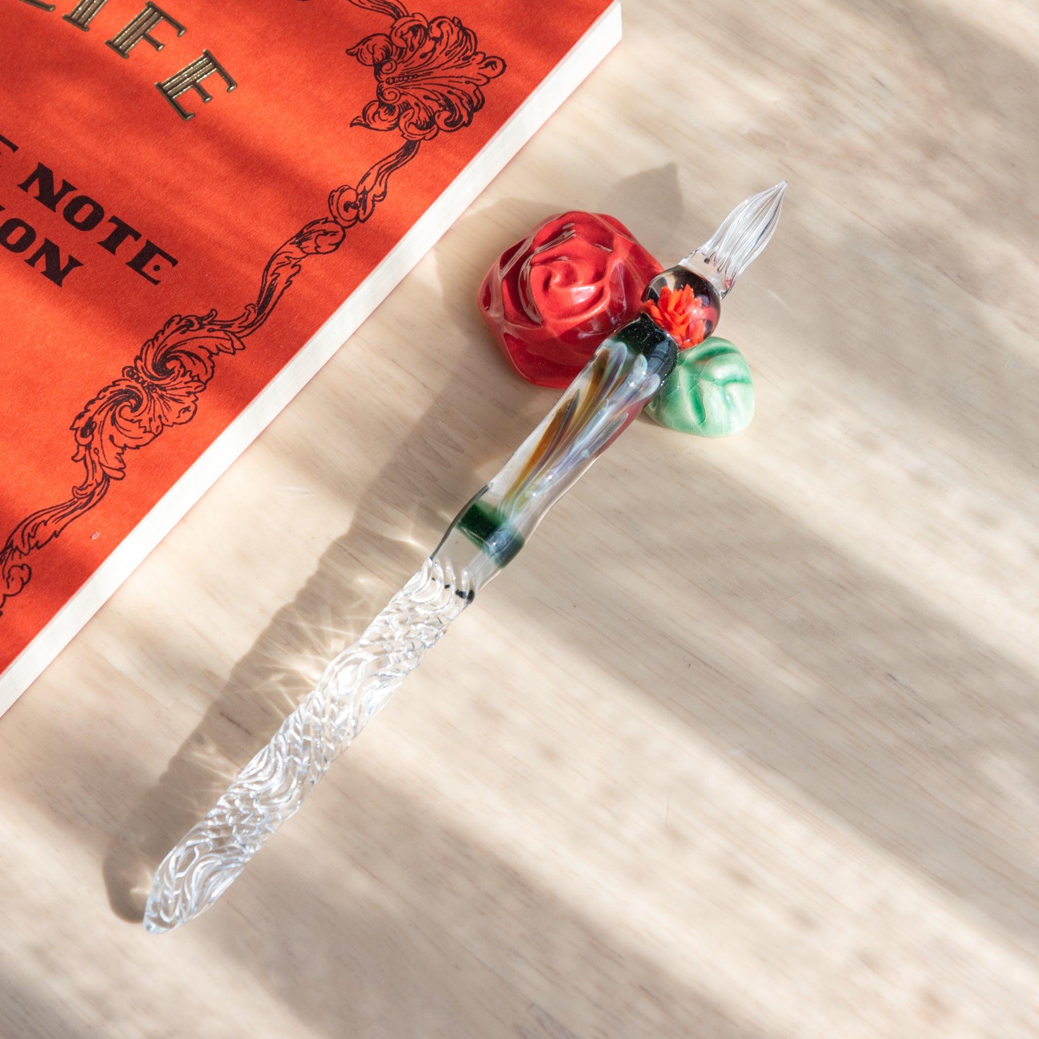 Handcrafted ceramic pen rest shaped like a red rose with a green leaf, paired with a decorative glass dip pen and Japanese stationery notebook.