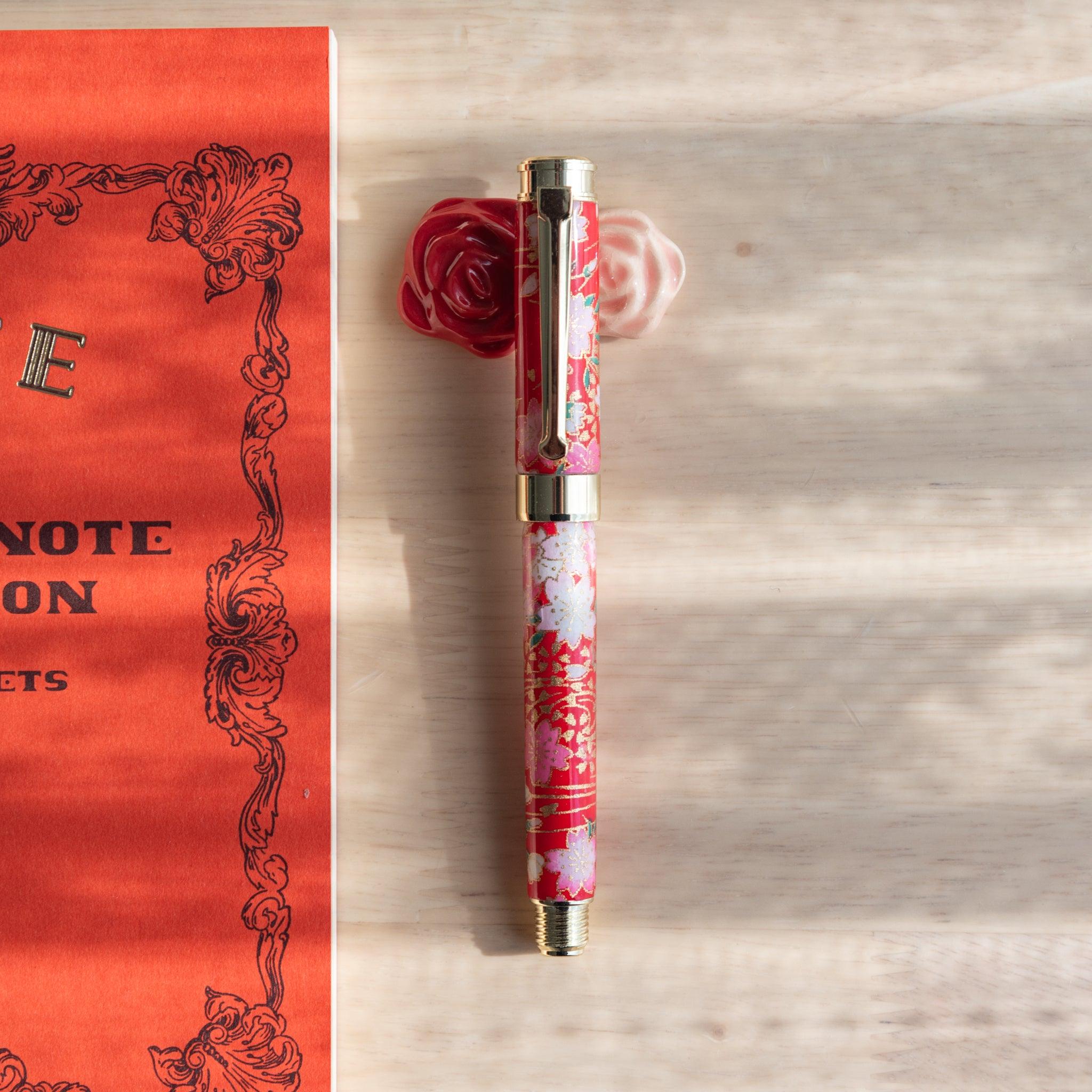 Floral Japanese fountain pen resting on a red and white rose-shaped ceramic pen rest, part of Japanese stationery, beside an orange notebook.