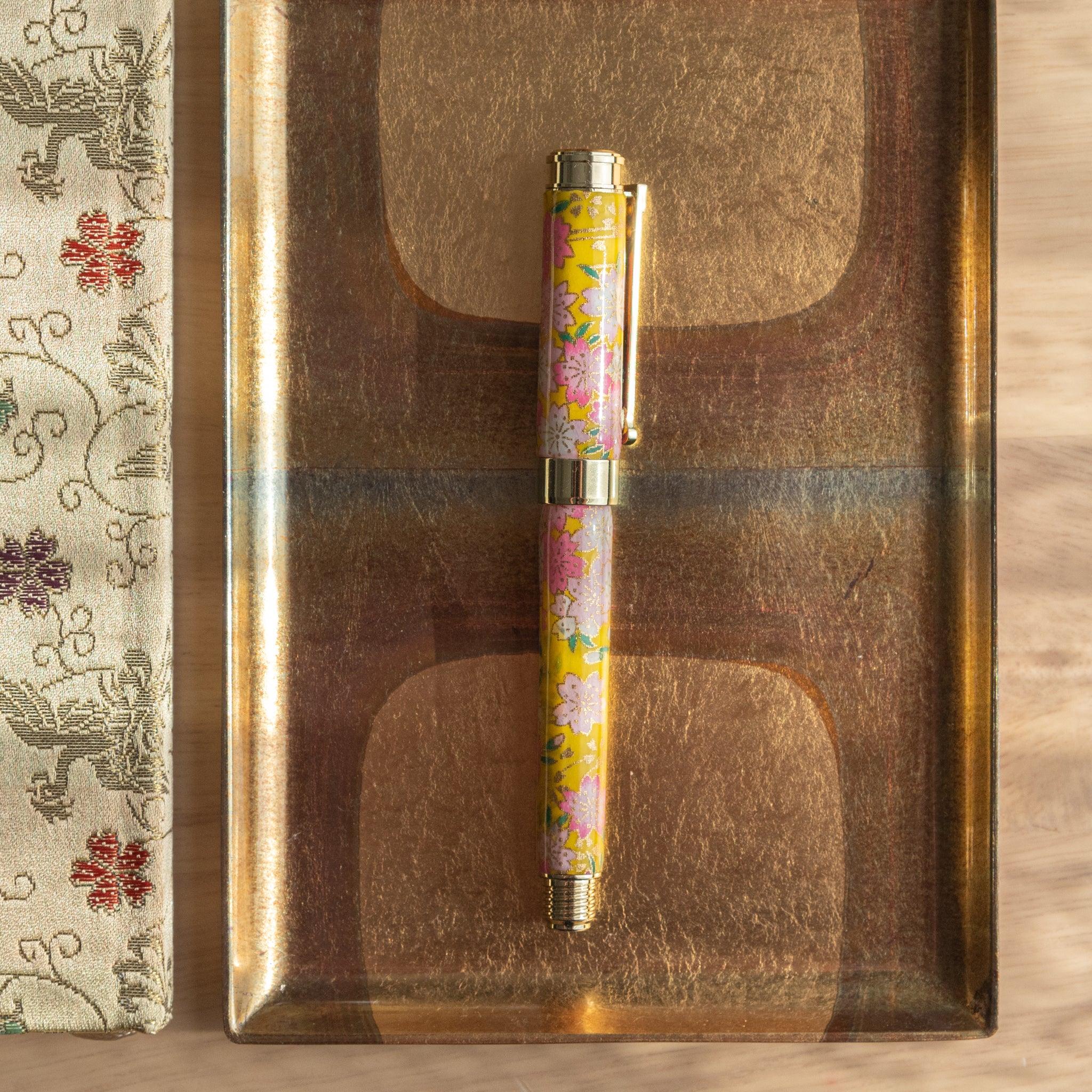 Closed Japanese fountain pen with floral sakura and canola design in yellow, resting on a golden tray. (84 characters)