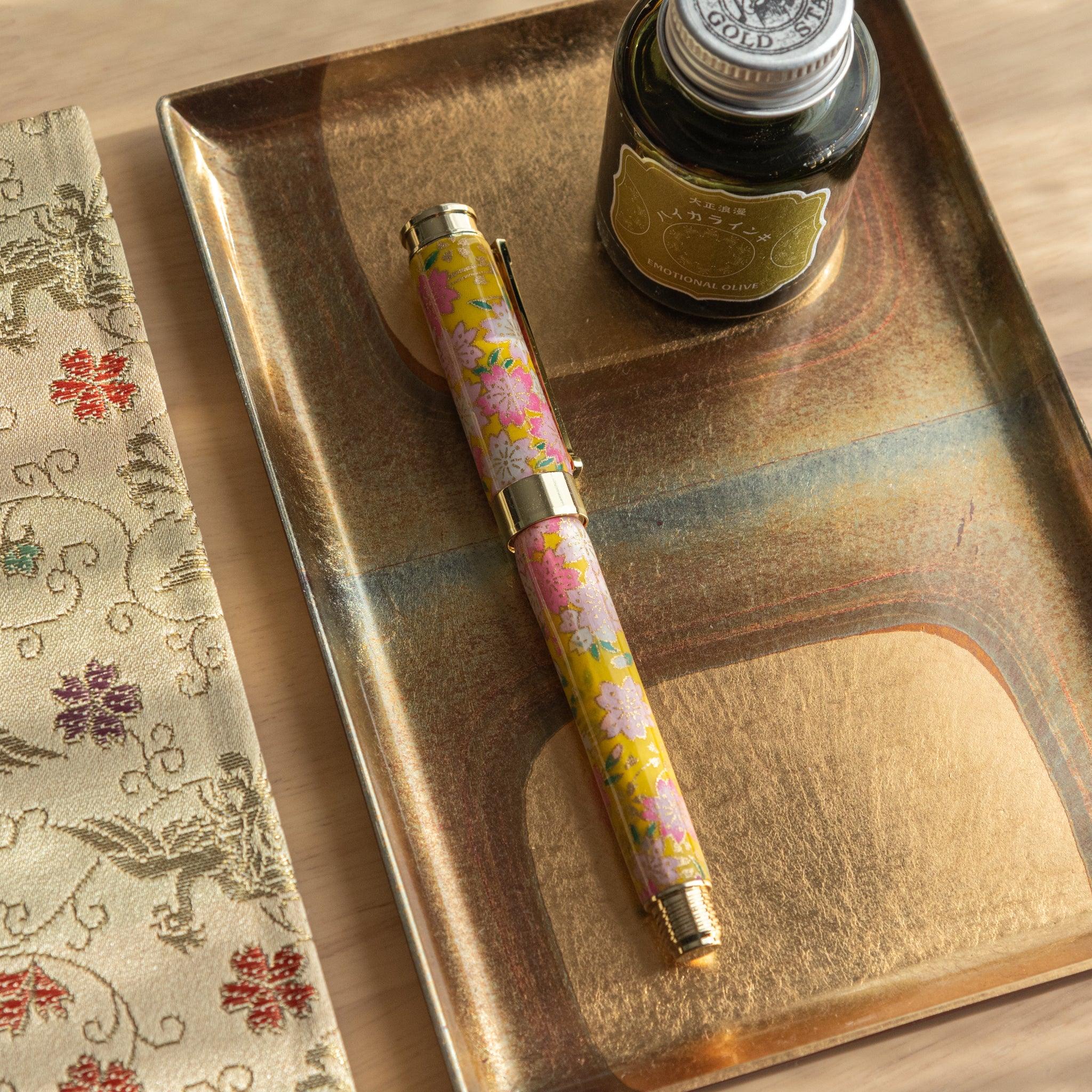 Closed Japanese fountain pen with sakura and canola pattern in pink and yellow, displayed with ink on a golden tray. (92 characters)