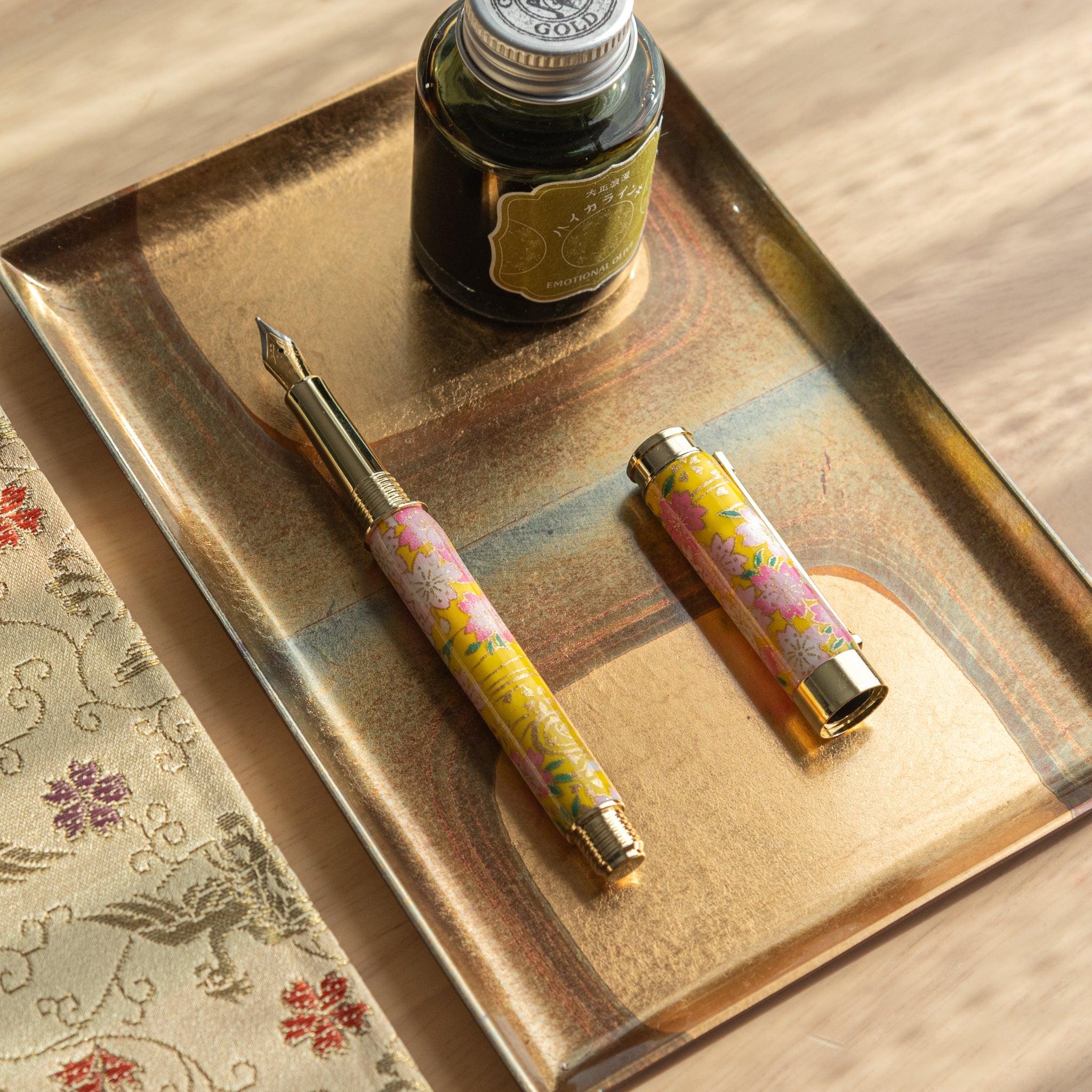 Open Japanese fountain pen with yellow sakura design on a golden tray, accompanied by a bottle of ink. (84 characters)