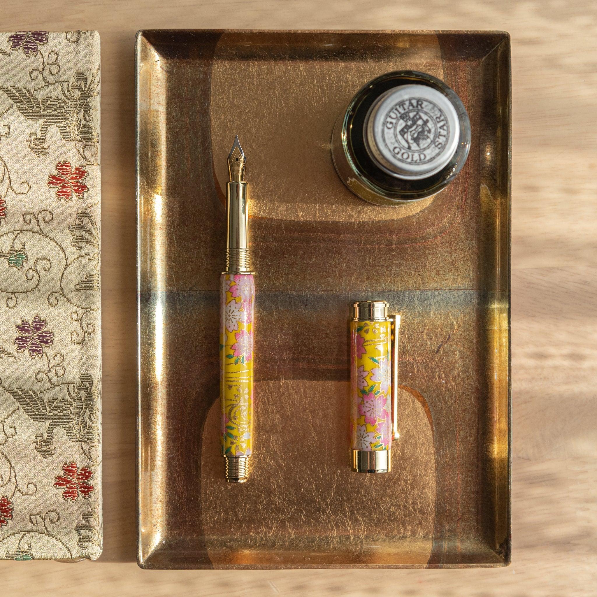 Japanese fountain pen featuring sakura and canola design in pink and yellow, displayed open and capped on a golden tray. (94 characters)