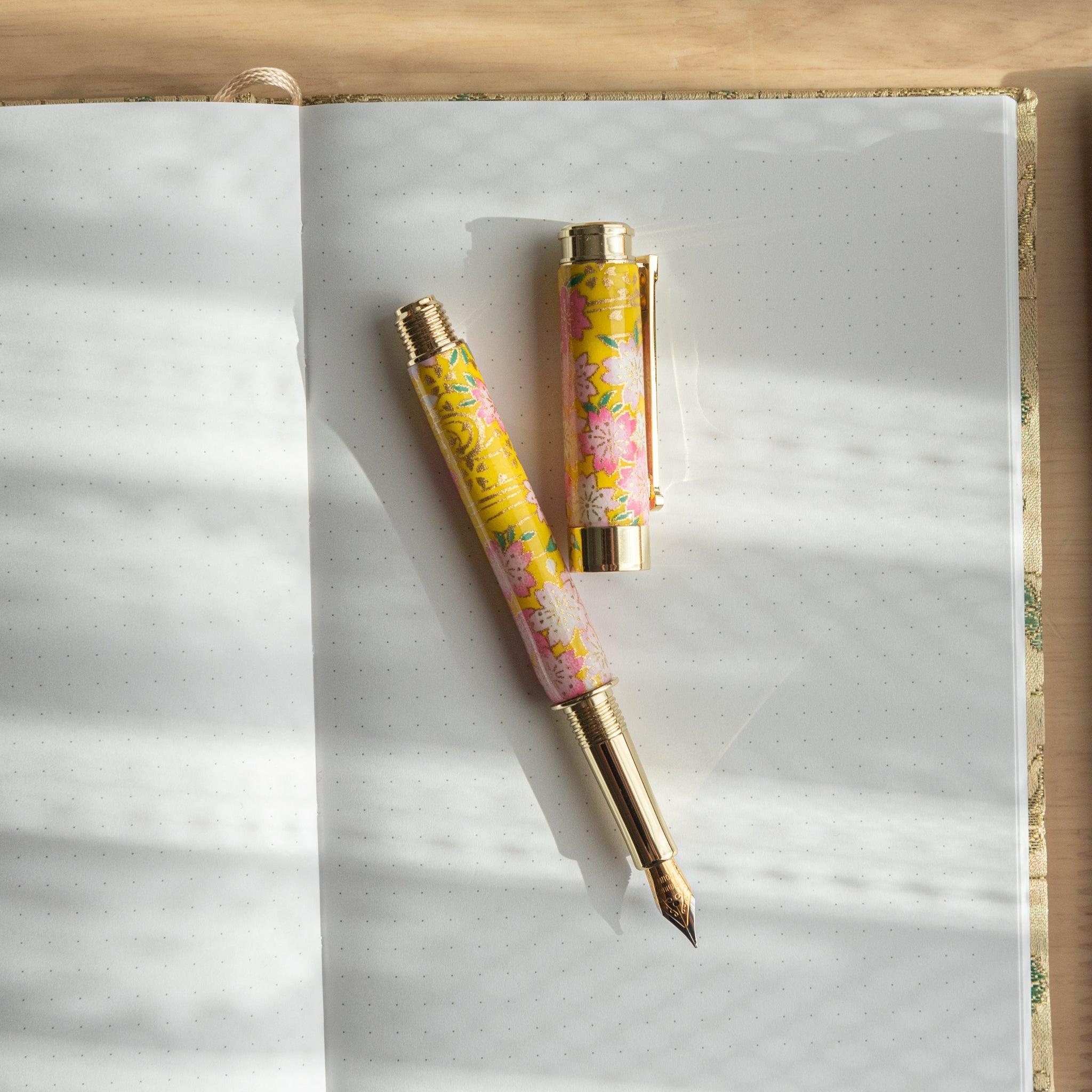 Japanese fountain pen with sakura and canola motif in yellow tones, resting open on a dotted notebook. (84 characters)