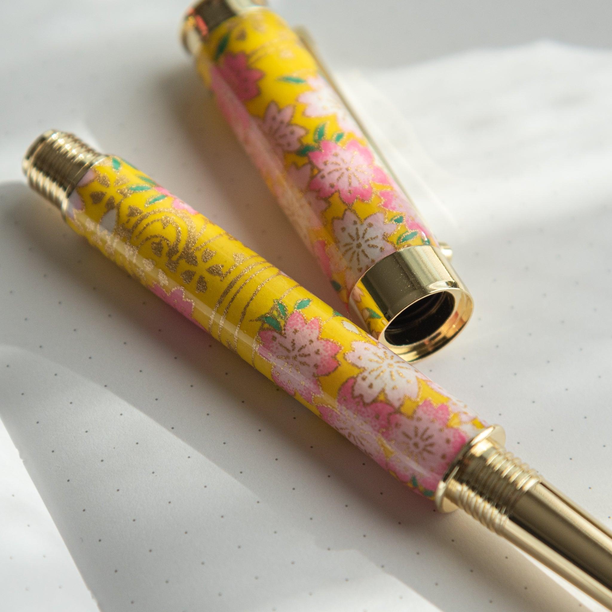 Close-up of Japanese fountain pen with sakura and canola motif in pink and yellow, placed on a dotted notebook. (92 characters)