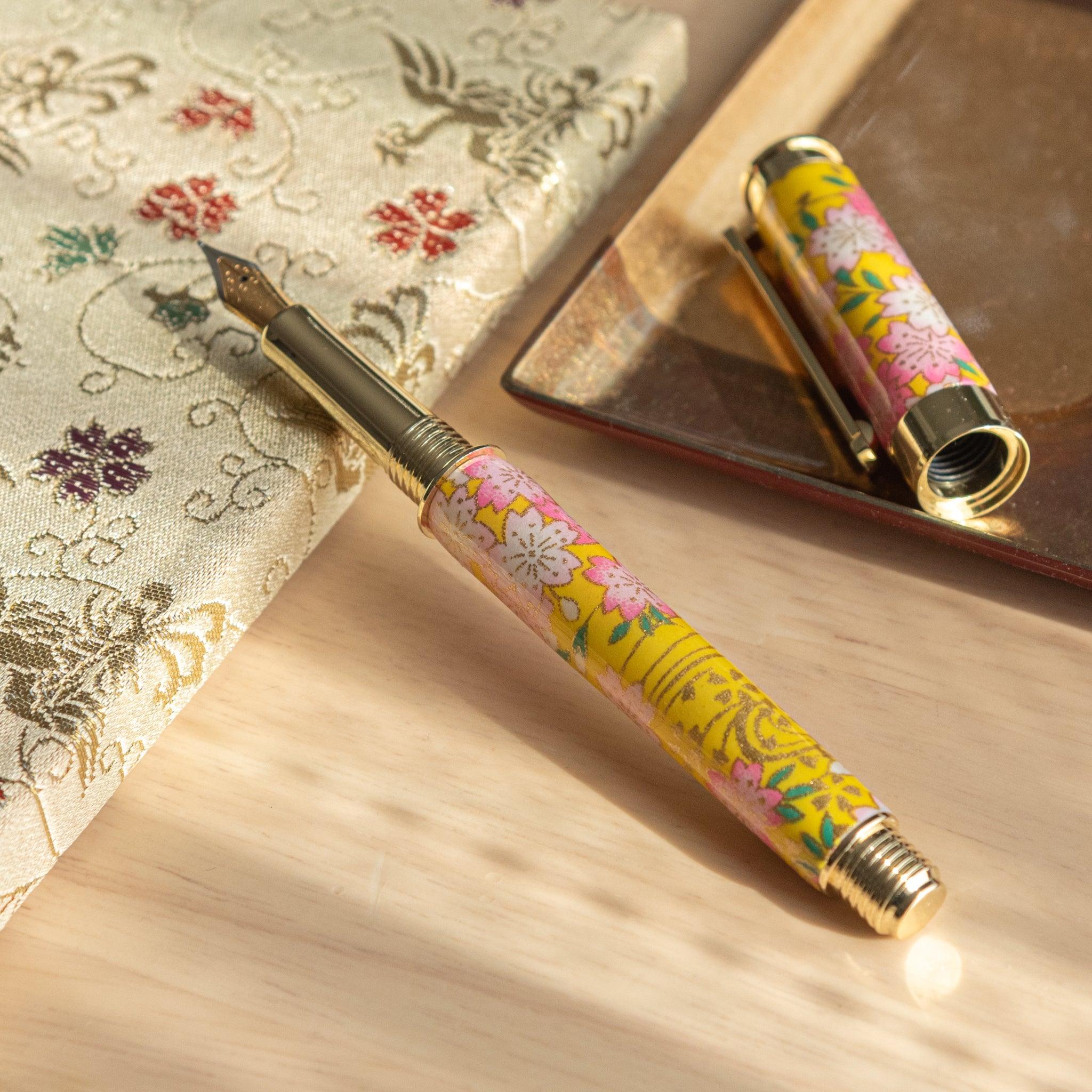 Open Japanese fountain pen with sakura and canola motif in yellow and pink, placed on a floral-patterned fabric. (92 characters)