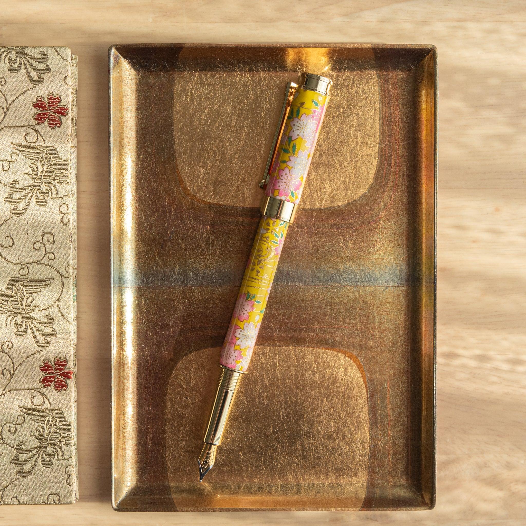 Open Japanese fountain pen with sakura and canola motif in yellow and pink, displayed on an elegant golden tray. (92 characters)