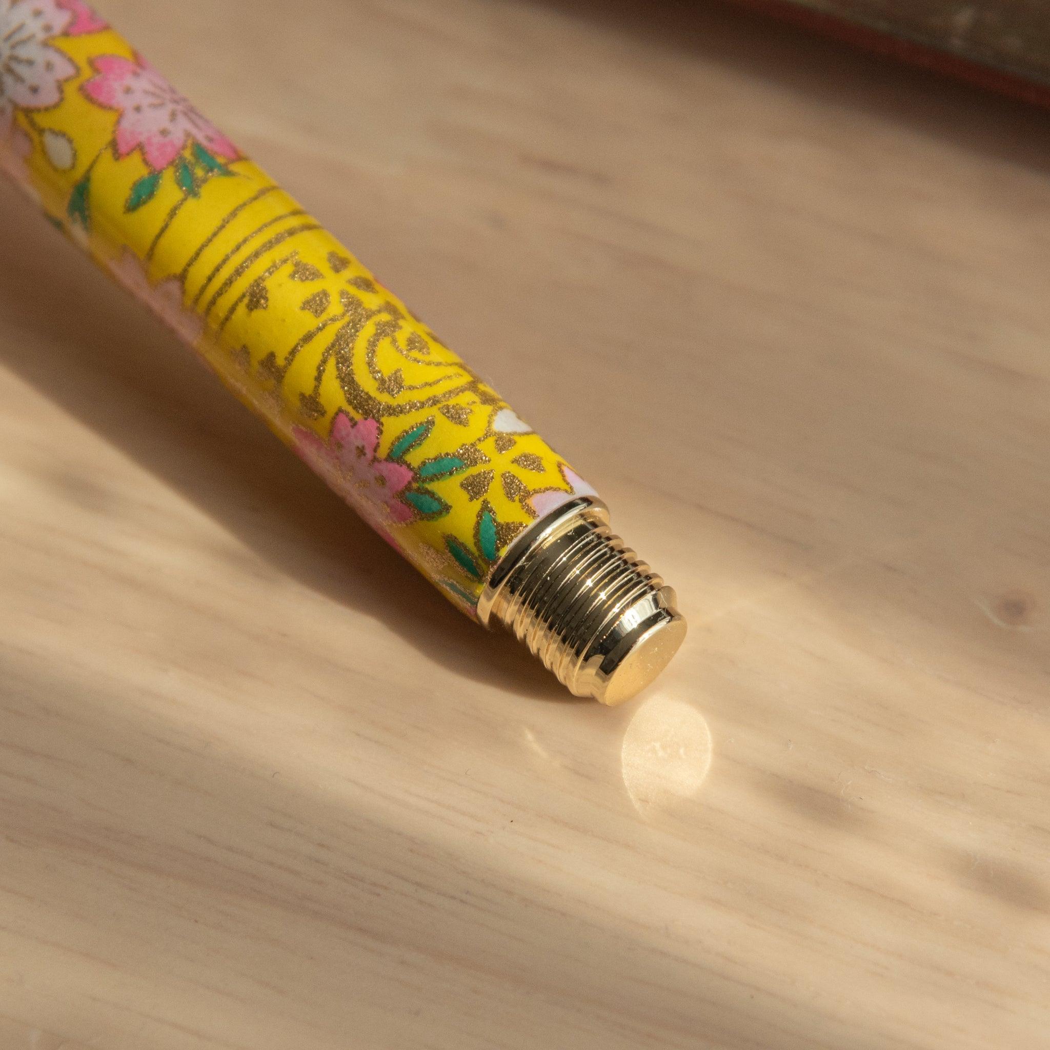 Close-up of Japanese fountain pen barrel with sakura and canola motif in yellow and pink, resting on a wooden surface. (92 characters)