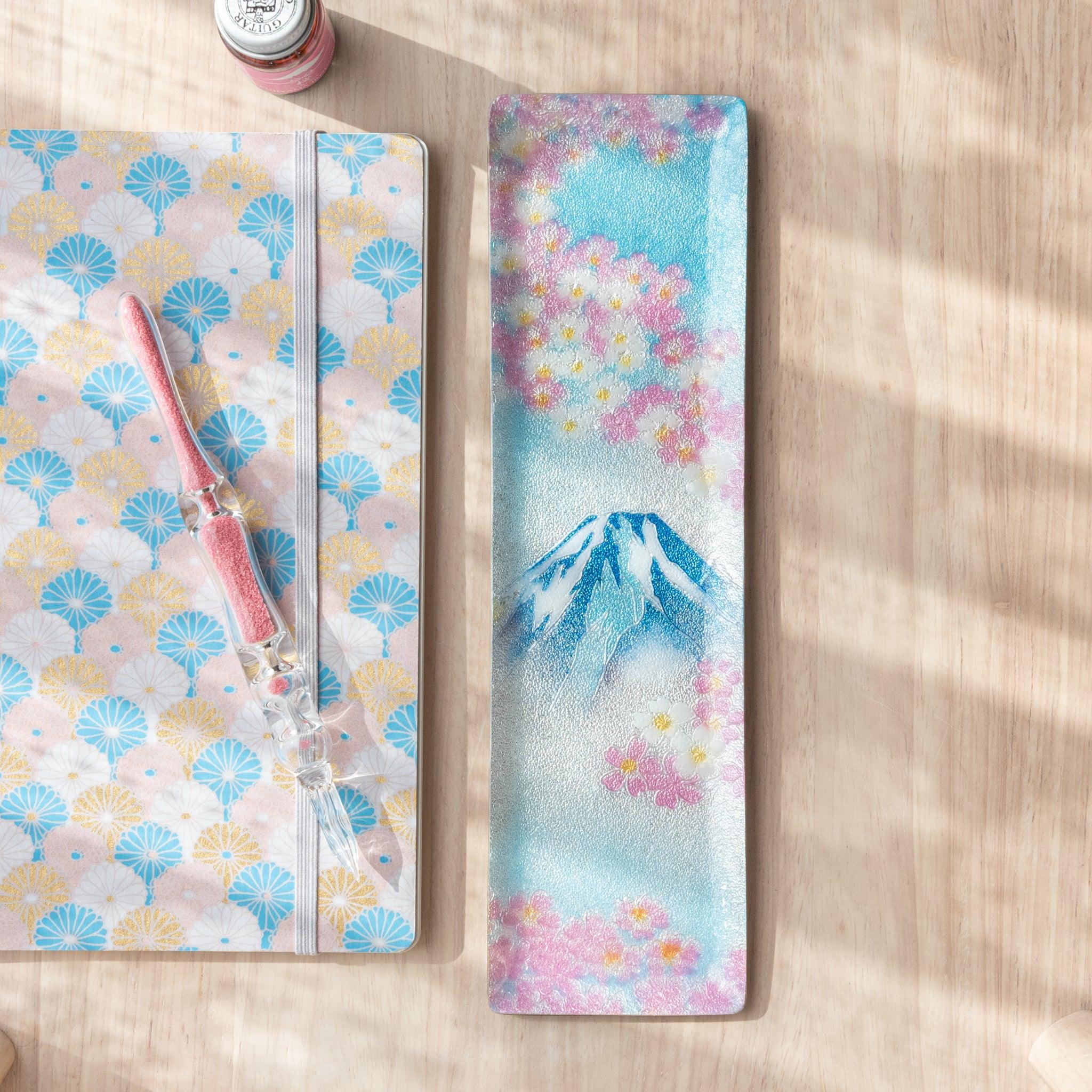 Rectangular tray with Mt. Fuji and cherry blossoms design, holding blue and pink glass pens, next to a fan-patterned notebook on a wooden surface.