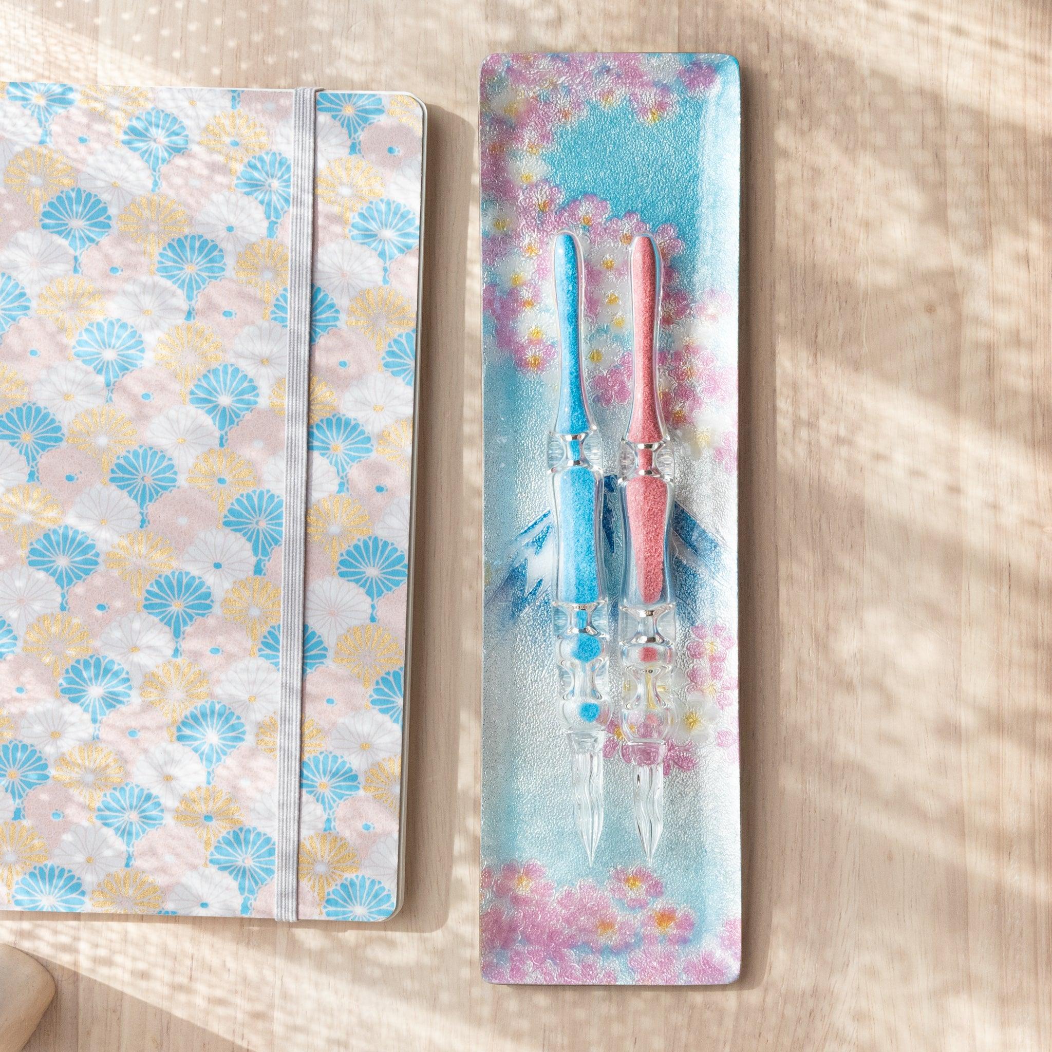 Angled view of Mt. Fuji tray adorned with pink cherry blossoms, placed beside a decorative notebook and pink glass pen on a light wooden background.