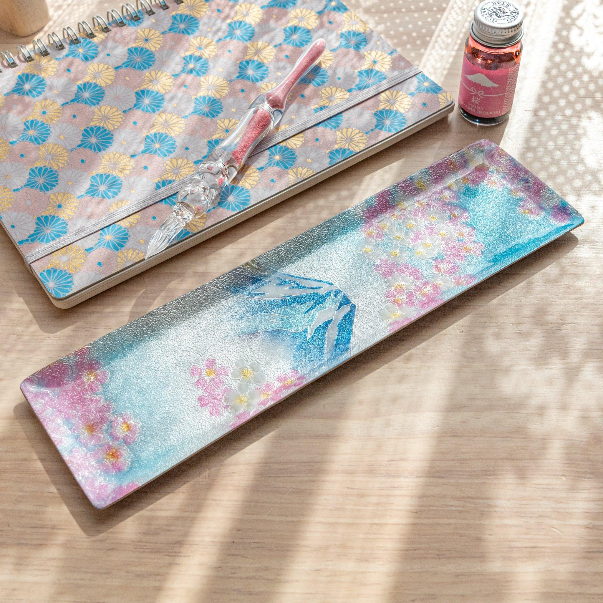 Rectangular tray with Mt. Fuji and sakura pattern, placed beside a pastel fan-patterned notebook and pink glass pen on a wooden surface.