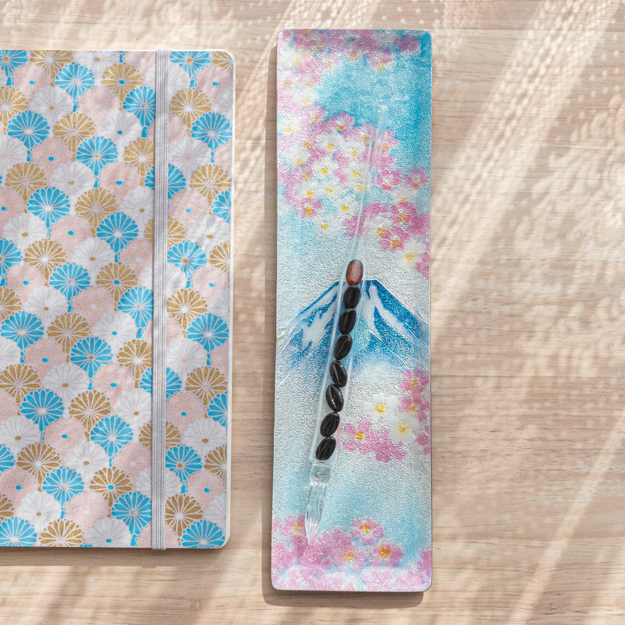 Rectangular tray with Mt. Fuji and cherry blossoms design, featuring a glass pen with black accents, next to a fan-patterned notebook on wood.