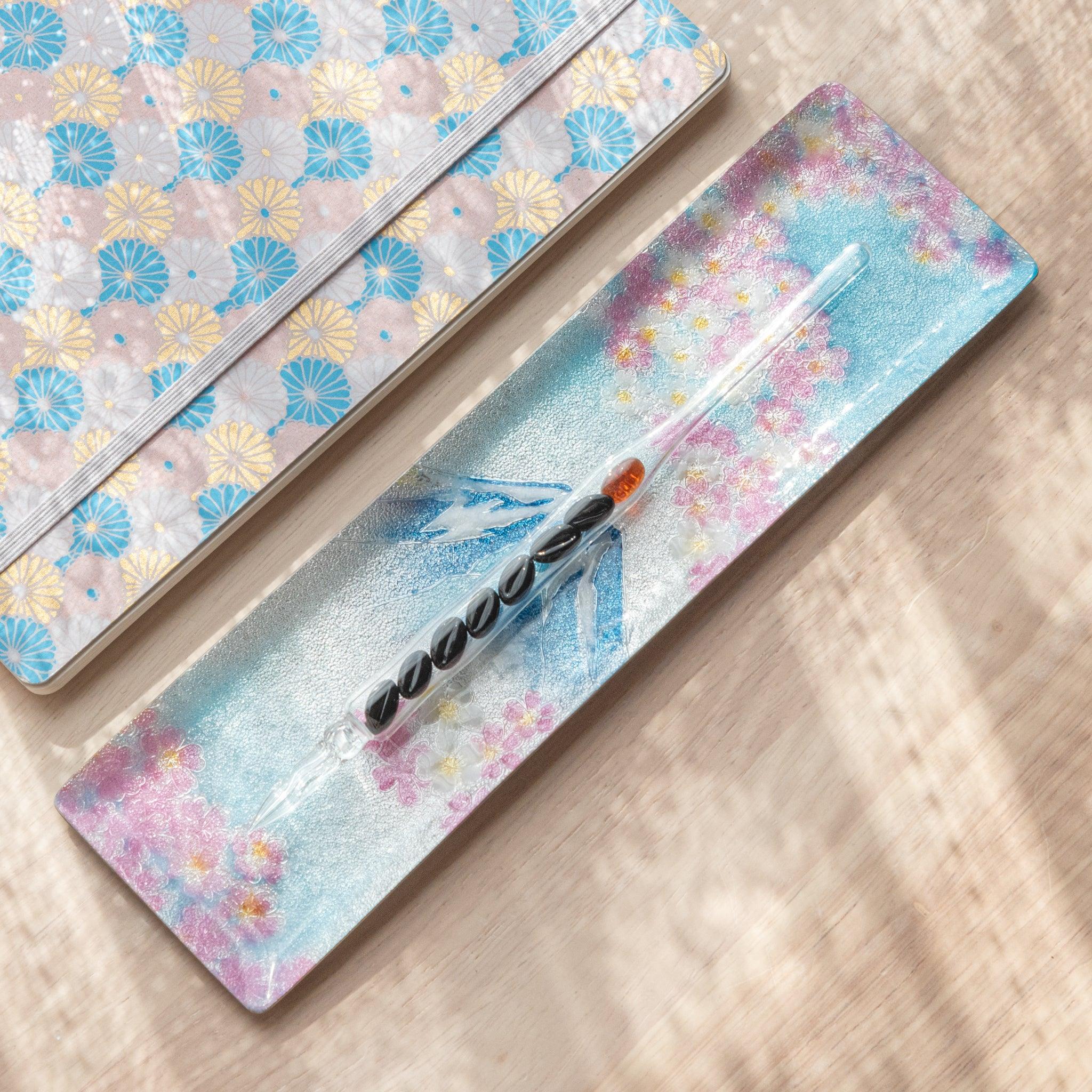 Angled view of Mt. Fuji tray adorned with pink cherry blossoms and glass pen, placed beside a decorative notebook on a wooden background.