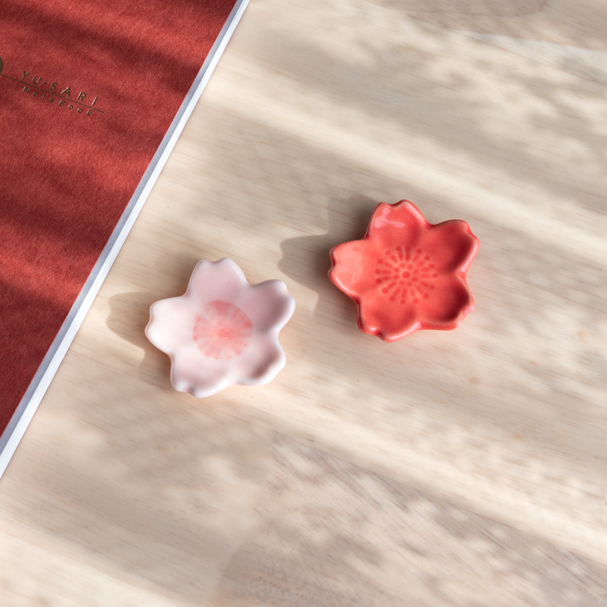 Two ceramic pen rests shaped like sakura flowers, in pink and red, part of Japanese stationery, placed on a wooden surface with an orange notebook.