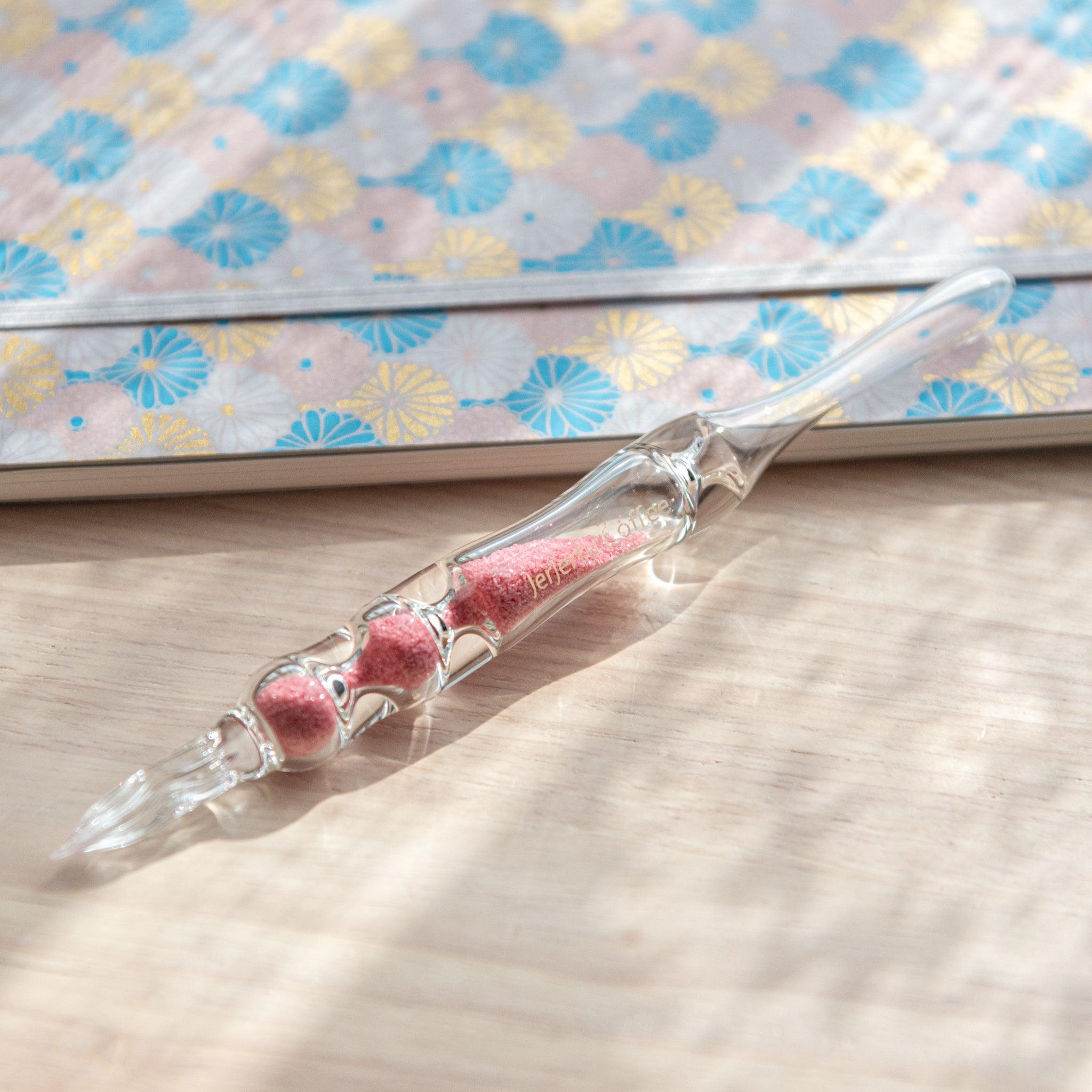 A glass dip pen with pink sand-like particles inside its shaft rests on a wooden surface beside a pastel floral-patterned notebook. The pen's transparent design highlights the vibrant pink particles inside.