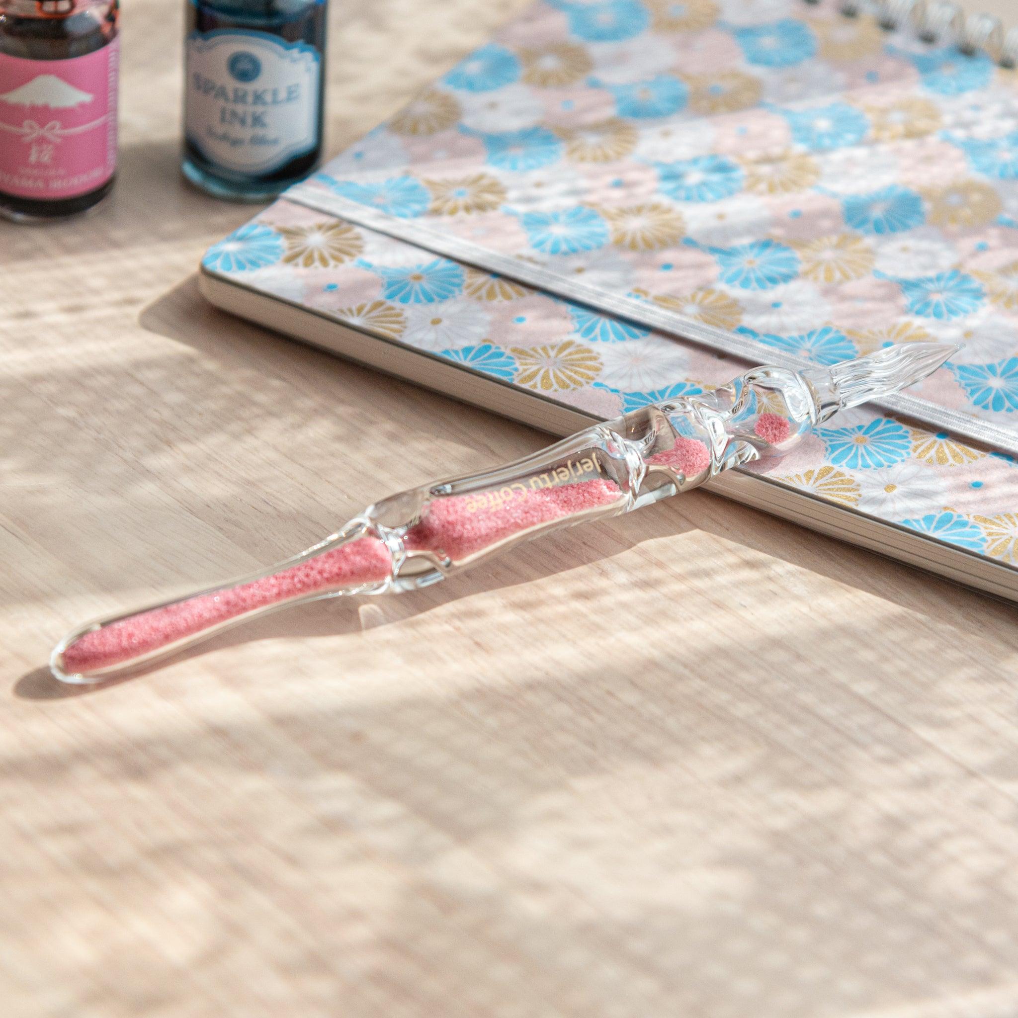 A glass dip pen with pink sand-like particles inside its shaft rests on a wooden surface beside a pastel floral-patterned notebook. The pen's transparent design highlights the vibrant pink particles inside.