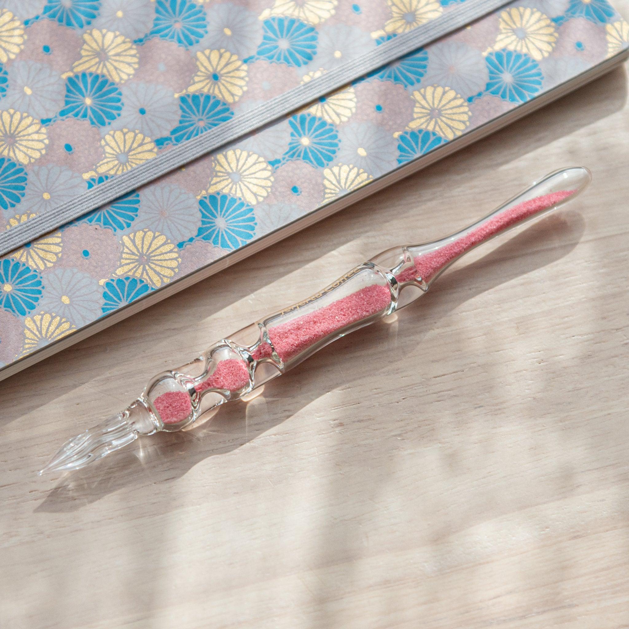 A glass dip pen with pink sand-like particles inside its shaft rests on a wooden surface beside a pastel floral-patterned notebook. The pen's transparent design highlights the vibrant pink particles inside.