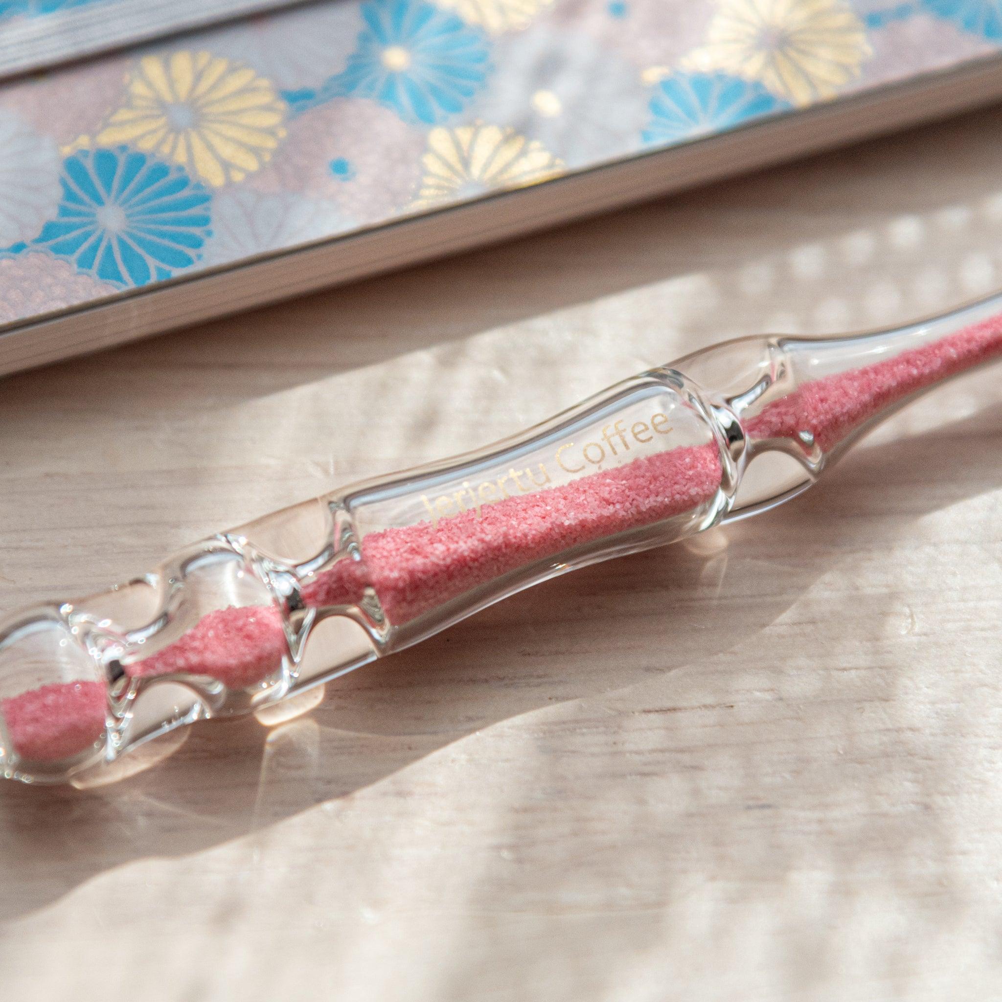 A glass dip pen with pink sand-like particles inside its shaft rests on a wooden surface beside a pastel floral-patterned notebook. The pen's transparent design highlights the vibrant pink particles inside.