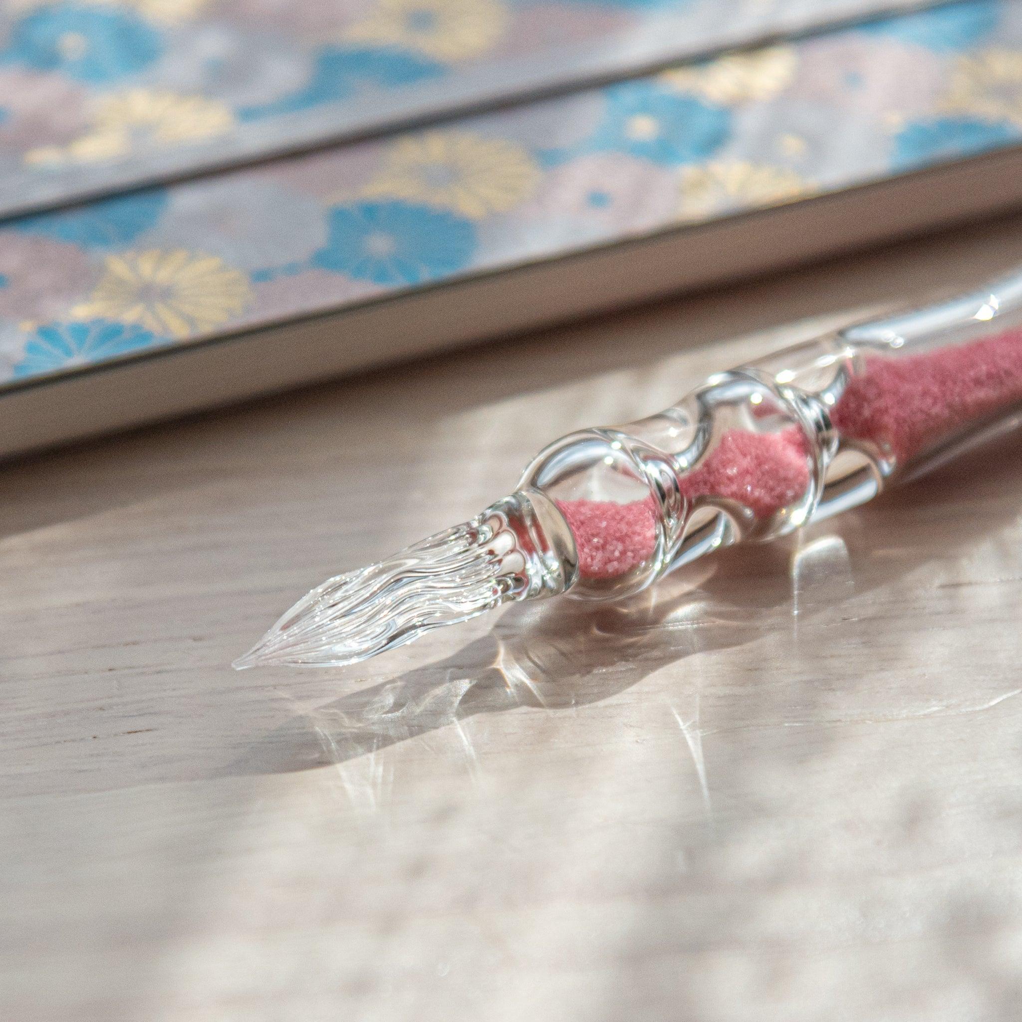 A glass dip pen with pink sand-like particles inside its shaft rests on a wooden surface beside a pastel floral-patterned notebook. The pen's transparent design highlights the vibrant pink particles inside.