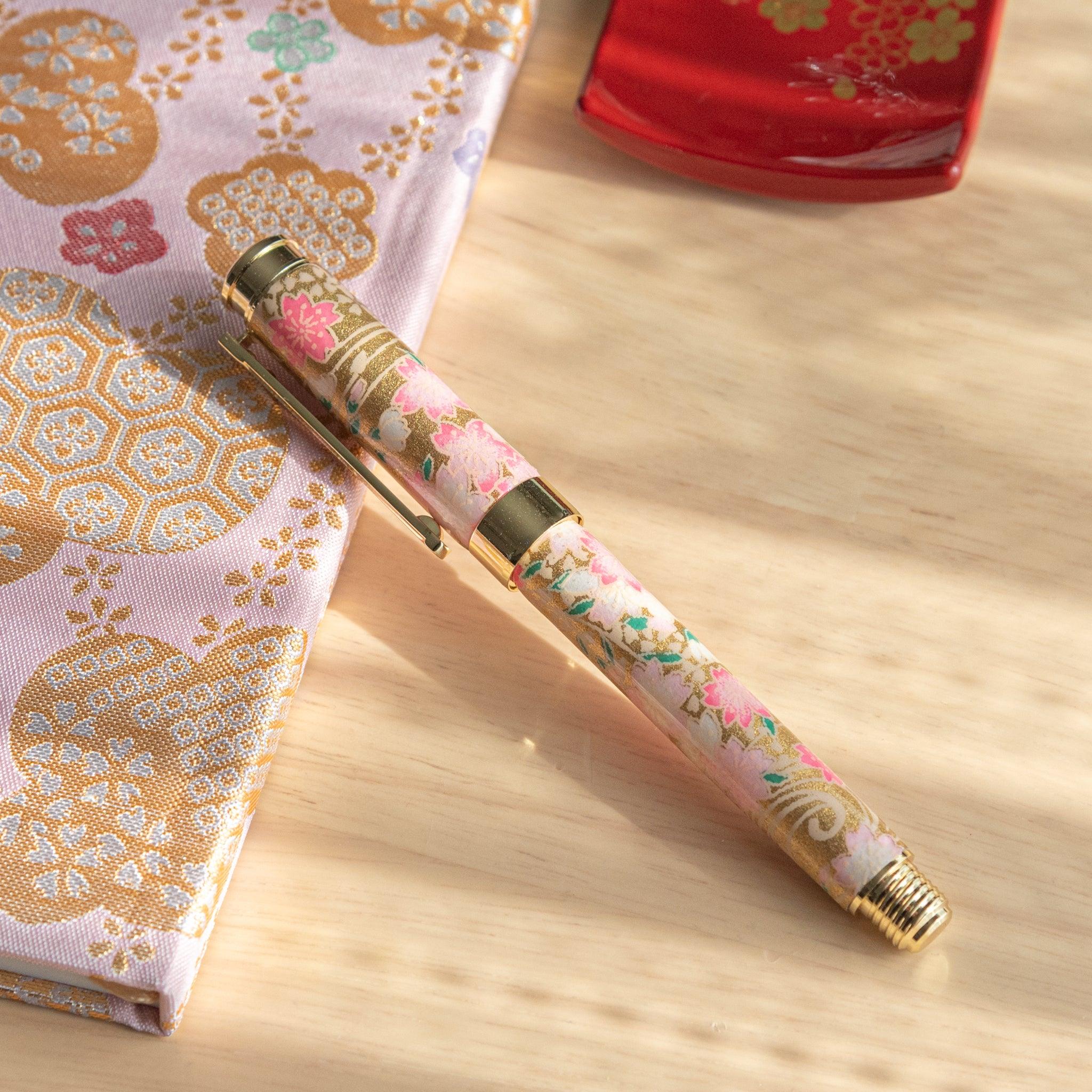 Pink and gold sakura-patterned fountain pen with intricate design, placed on a matching patterned fabric. (97 characters)