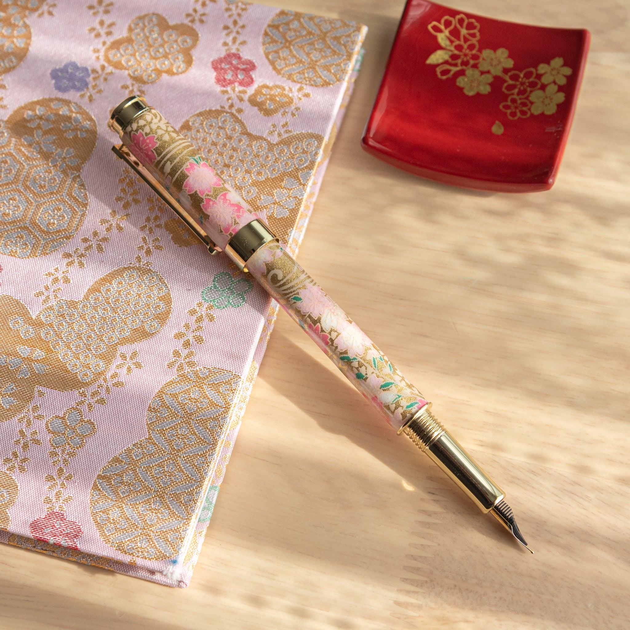 Open pink and gold fountain pen with sakura motif, displayed on a golden floral-patterned fabric. (91 characters)