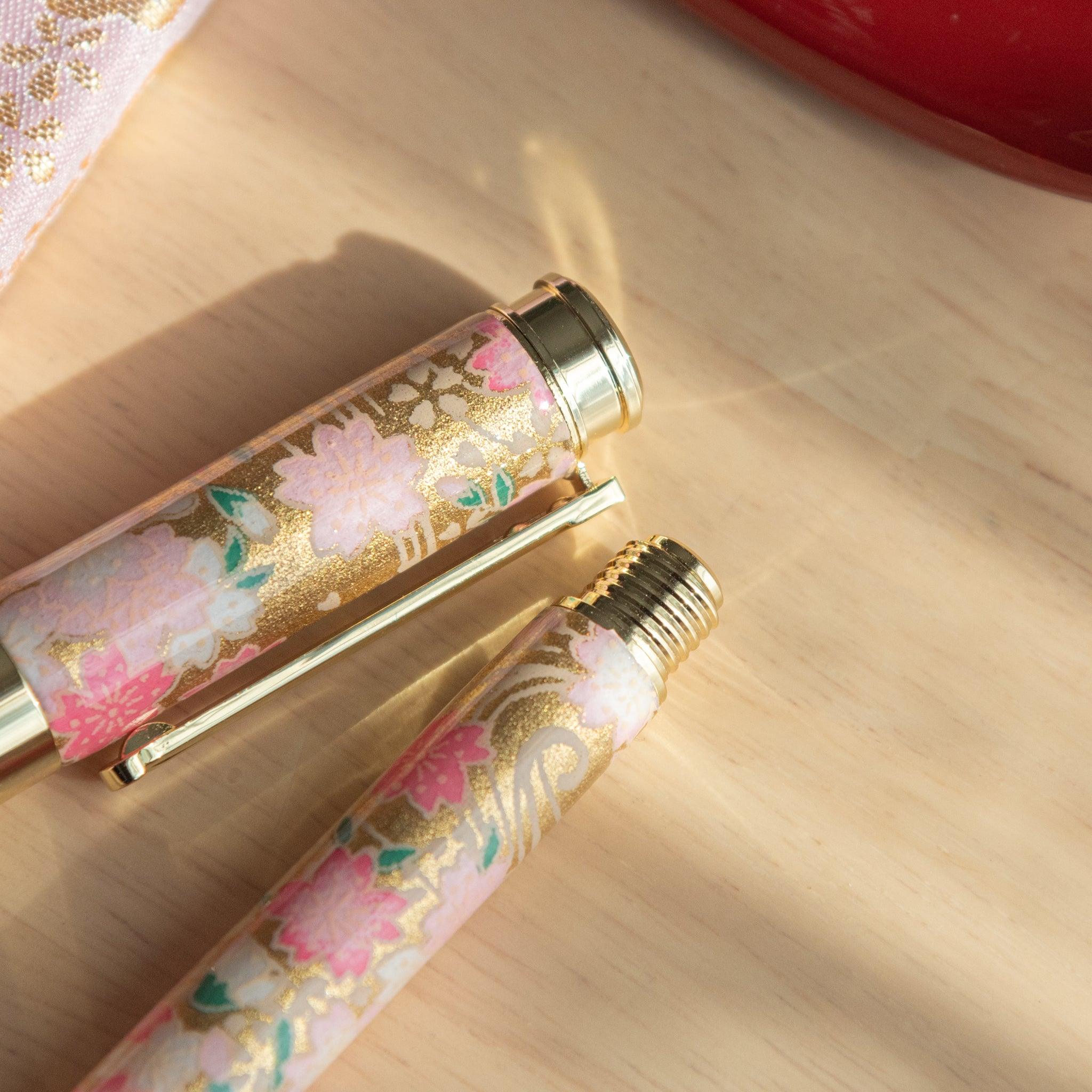 Close-up of a sakura-themed fountain pen cap and body with intricate gold detailing on a light wooden surface. (99 characters)