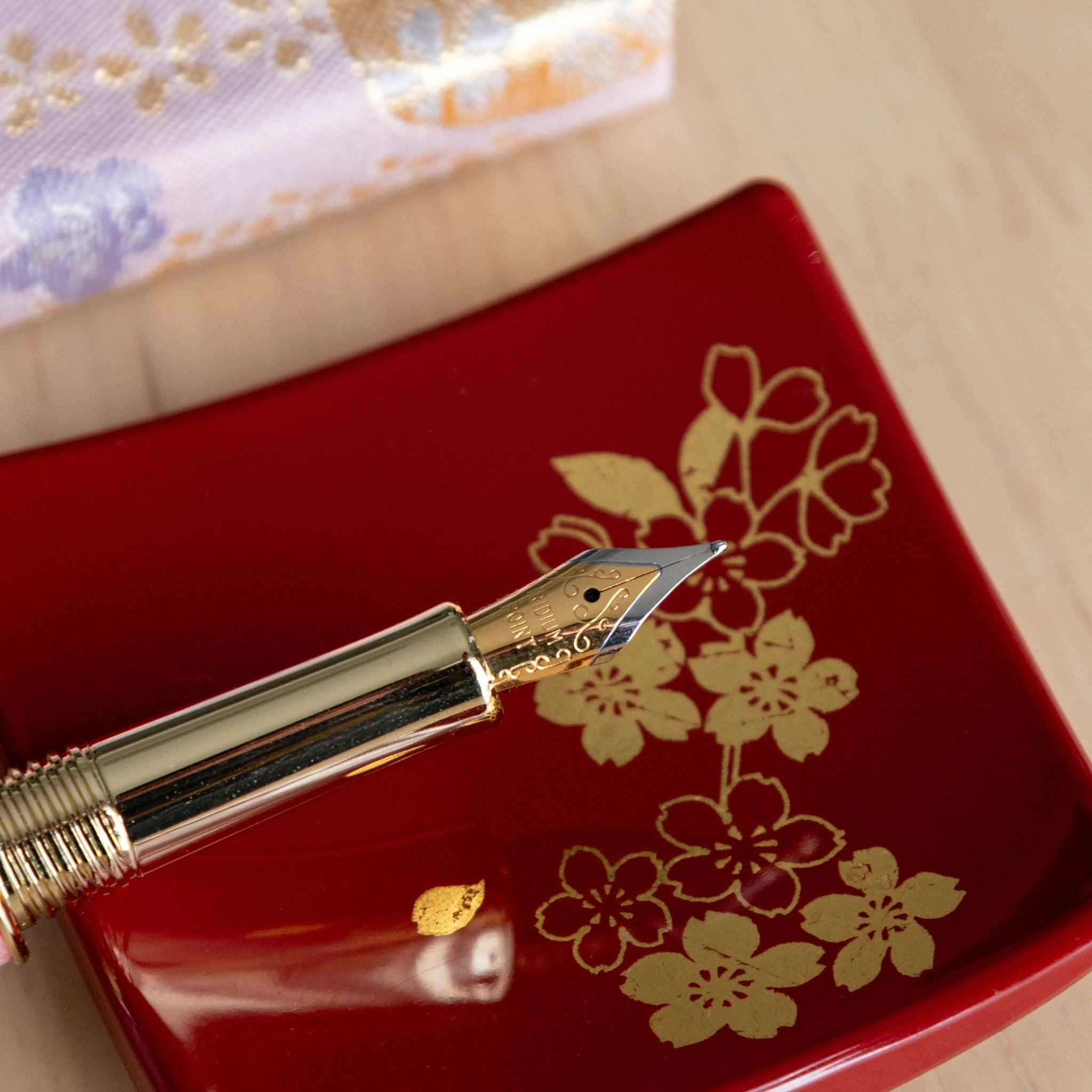 Gold-nibbed sakura-themed fountain pen resting on a red tray with golden floral accents, close-up view. (92 characters)