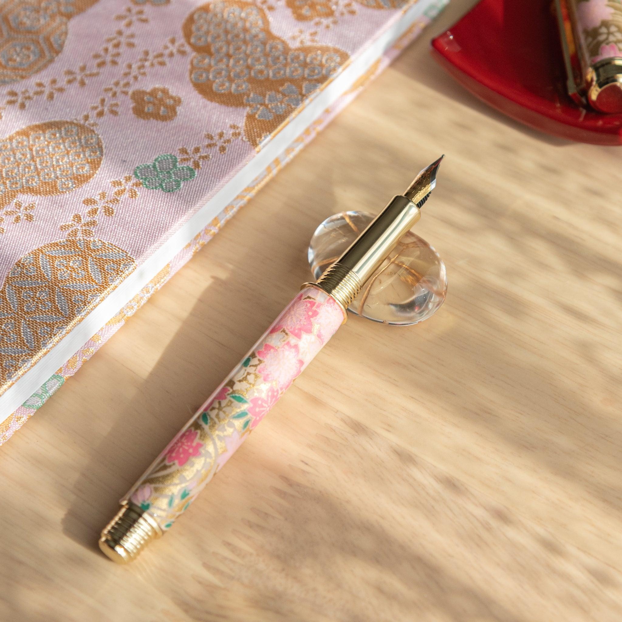 Open pink and gold sakura-patterned fountain pen resting on a clear glass stand next to golden-patterned fabric. (100 characters)