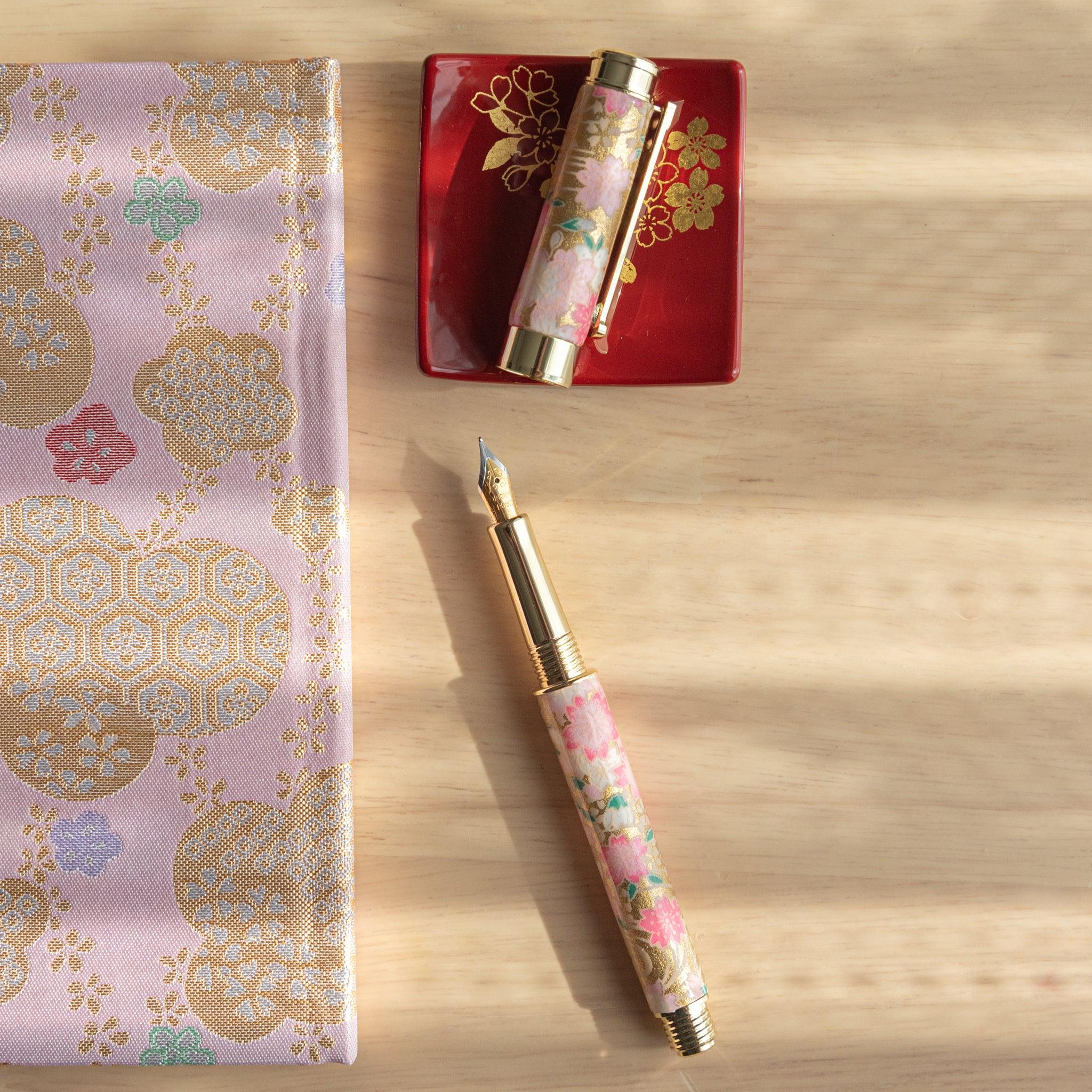 Open pink sakura-patterned fountain pen with gold details, displayed with cap on a red tray and golden-patterned fabric. (103 characters)