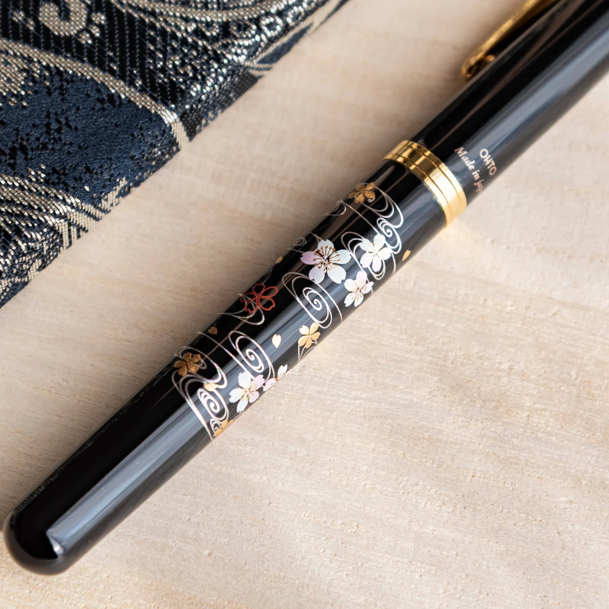 Black Yamanaka fountain pen with gold and sakura river motifs placed on a desk beside a black patterned fabric note.