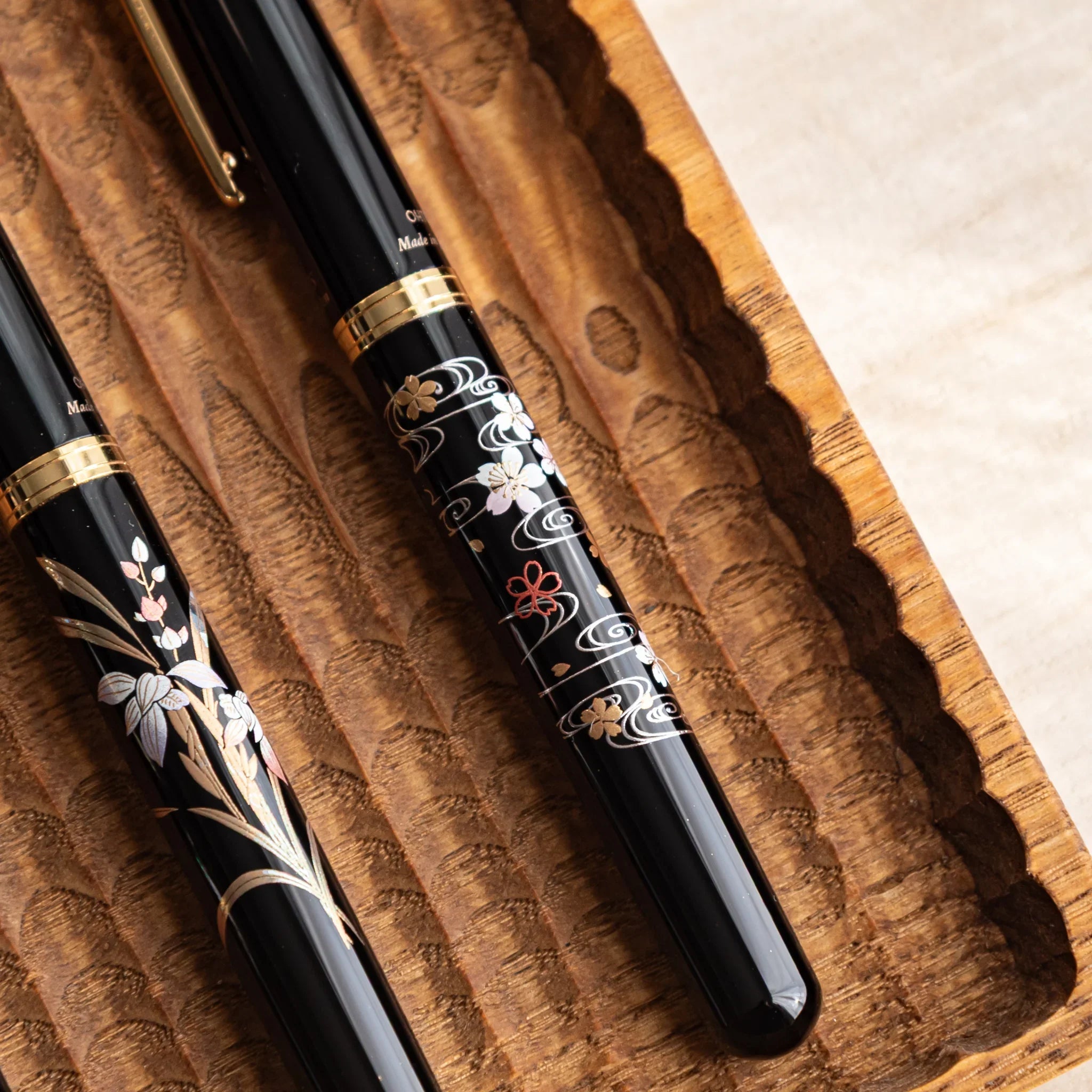 Two black Yamanaka fountain pens with gold sakura and river motifs resting in a carved wooden tray on a light desk.