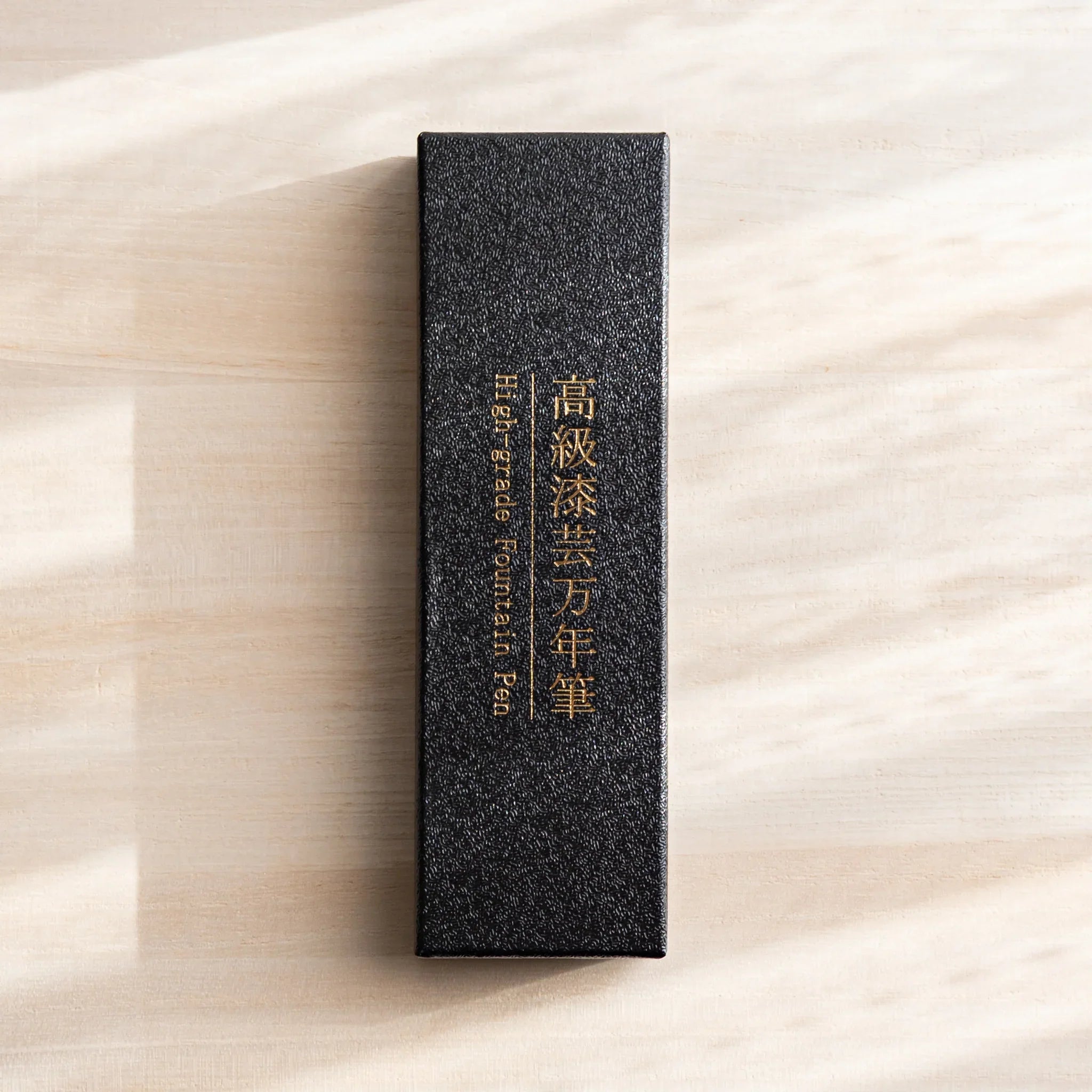 Black textured gift box with gold Japanese lettering for a Yamanaka fountain pen placed on a light wood desk.