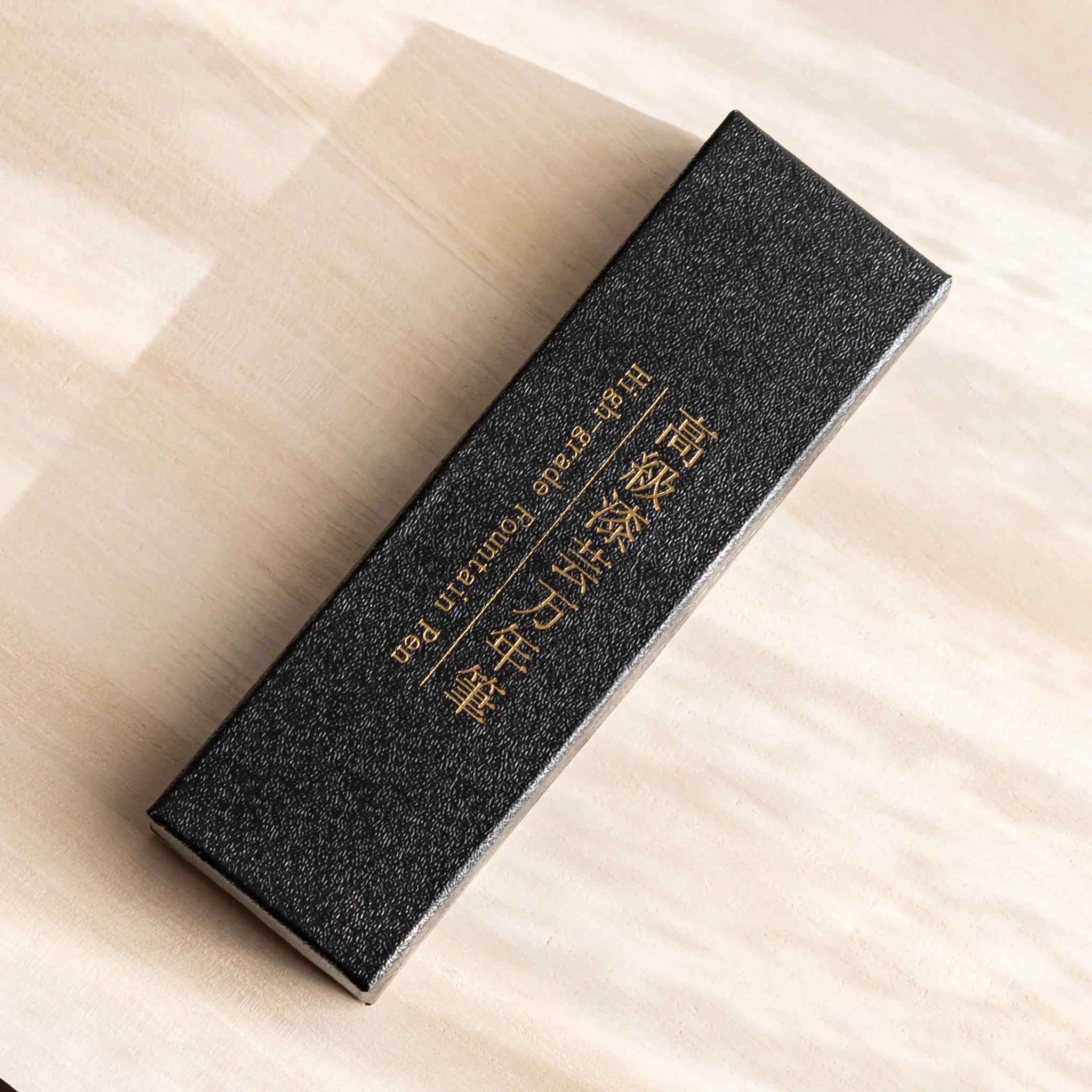 Black textured gift box with gold Japanese lettering for a Yamanaka fountain pen, placed diagonally on a wooden desk.