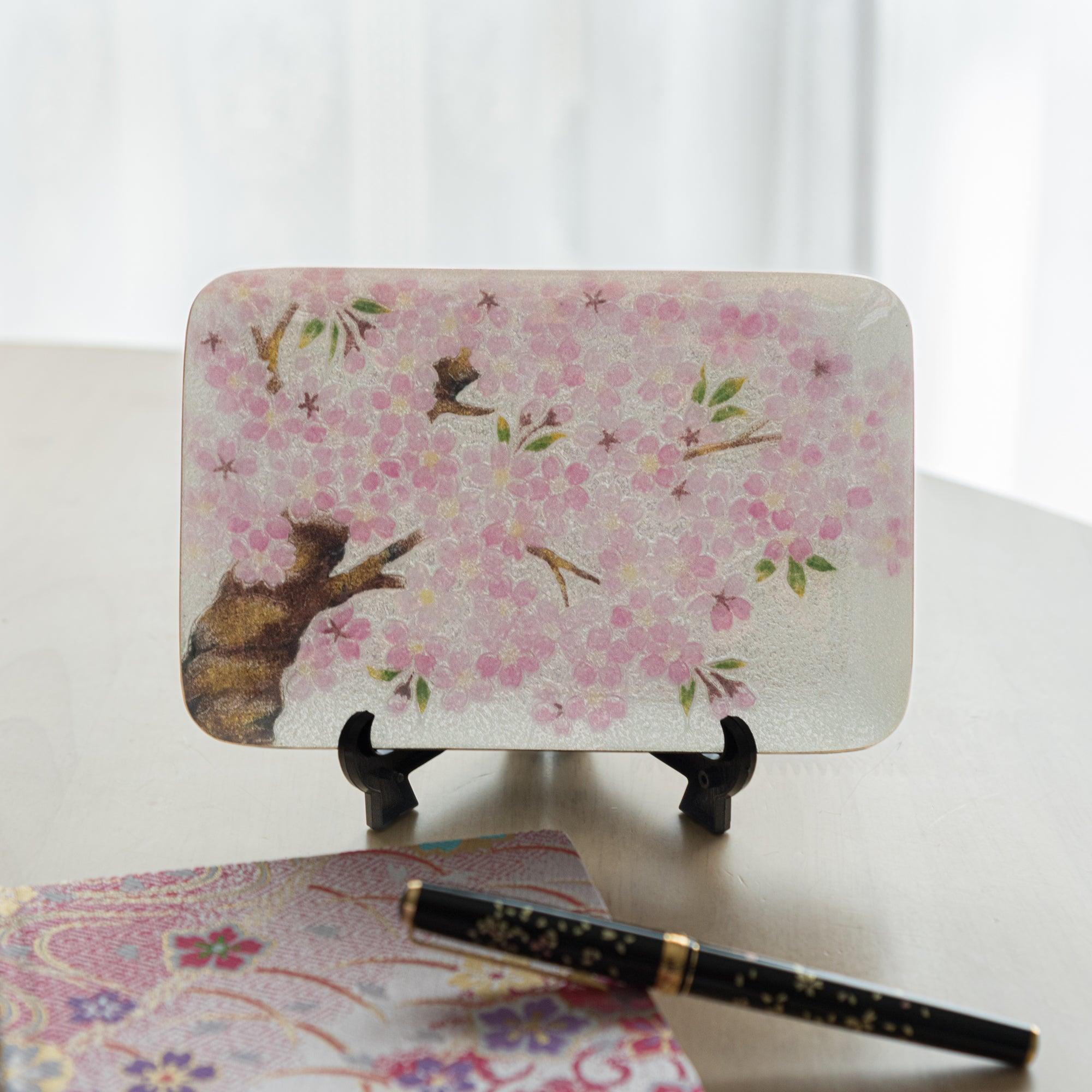 Decorative tray with cherry blossoms presented upright on a black stand, paired with a floral-patterned notebook and pen.  