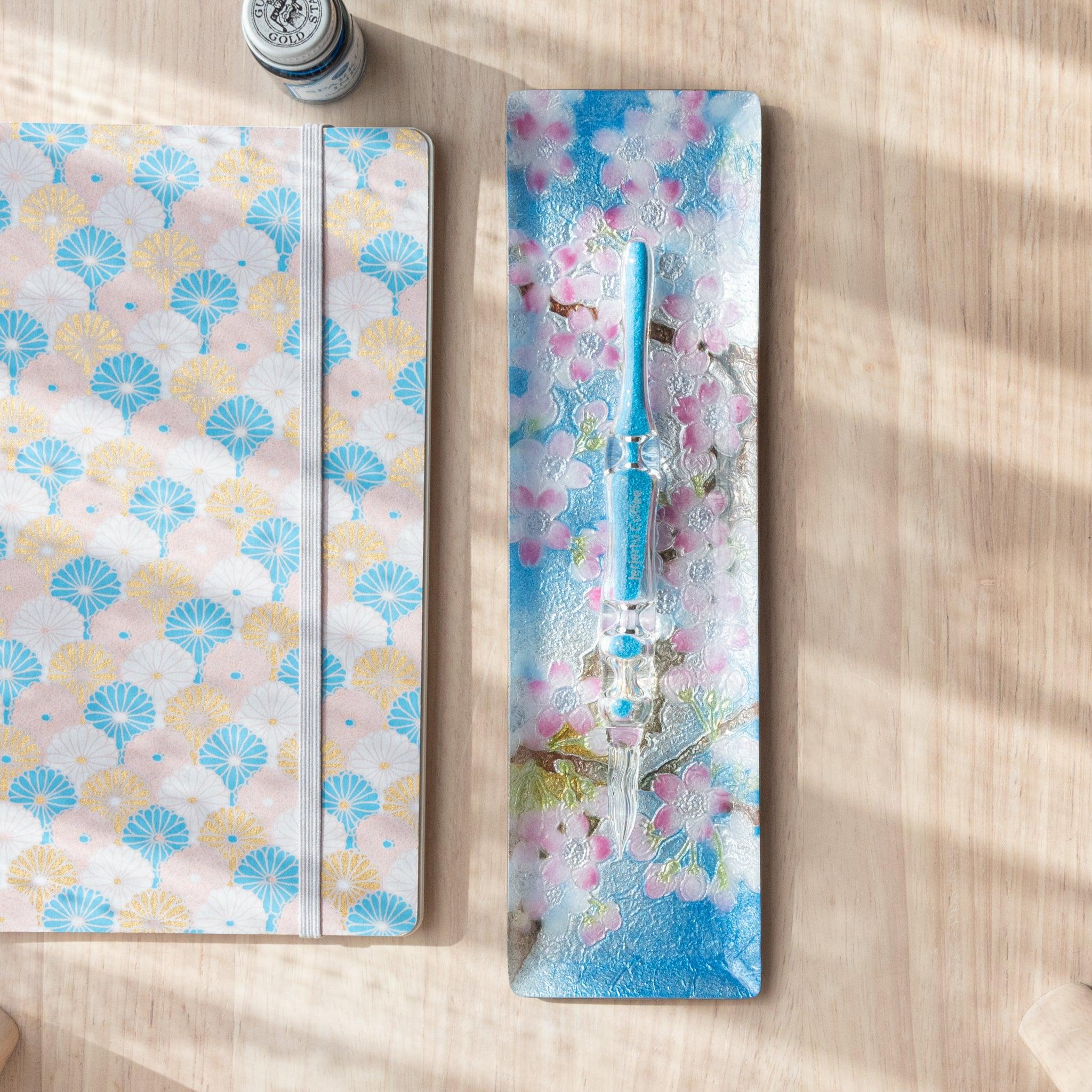 Rectangular tray with cherry blossom pattern in pink and white, holding a blue glass pen, beside a fan-patterned notebook on a wooden surface.