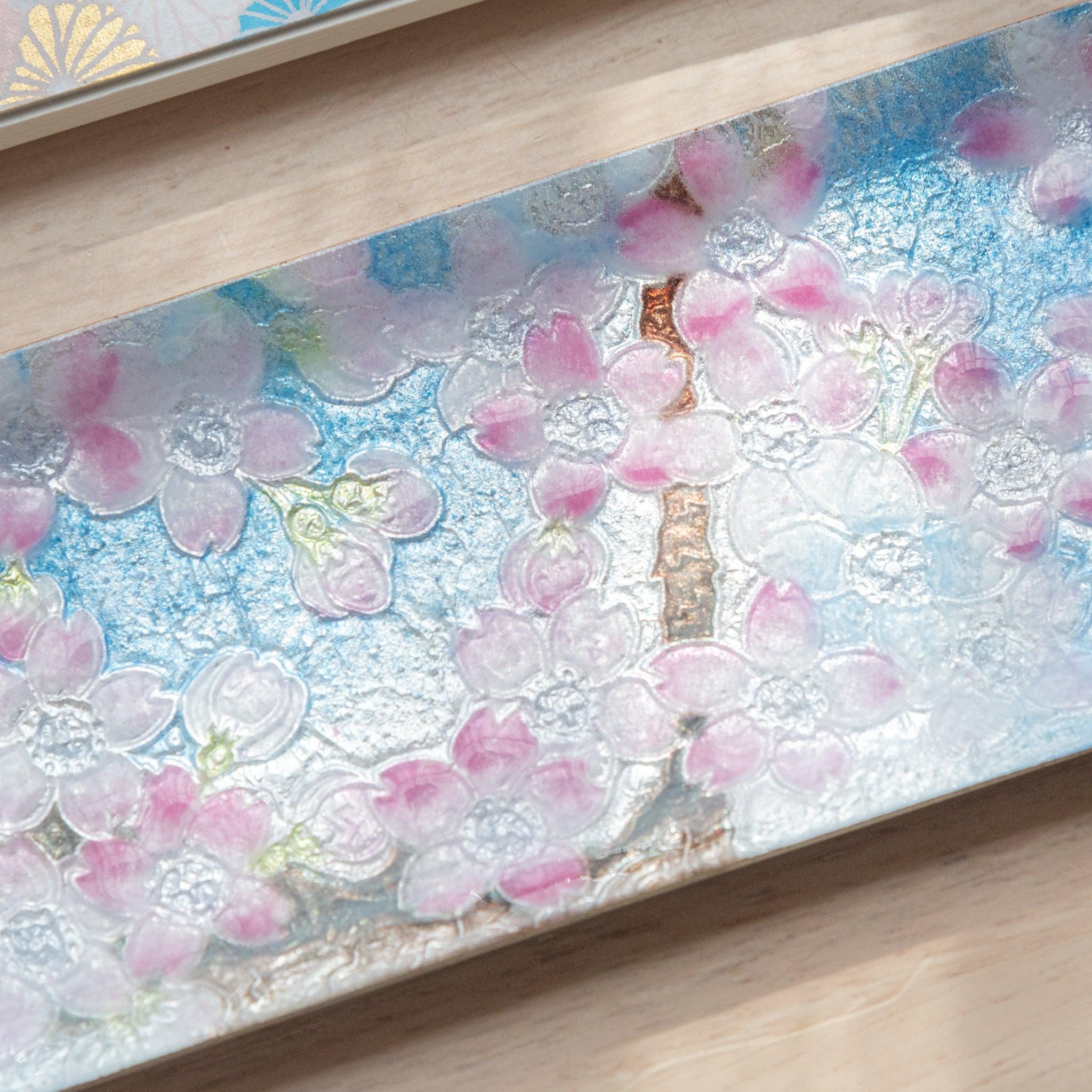 Rectangular tray adorned with pink cherry blossoms, placed next to a fan-patterned notebook and blue glass pen on a light wood background.