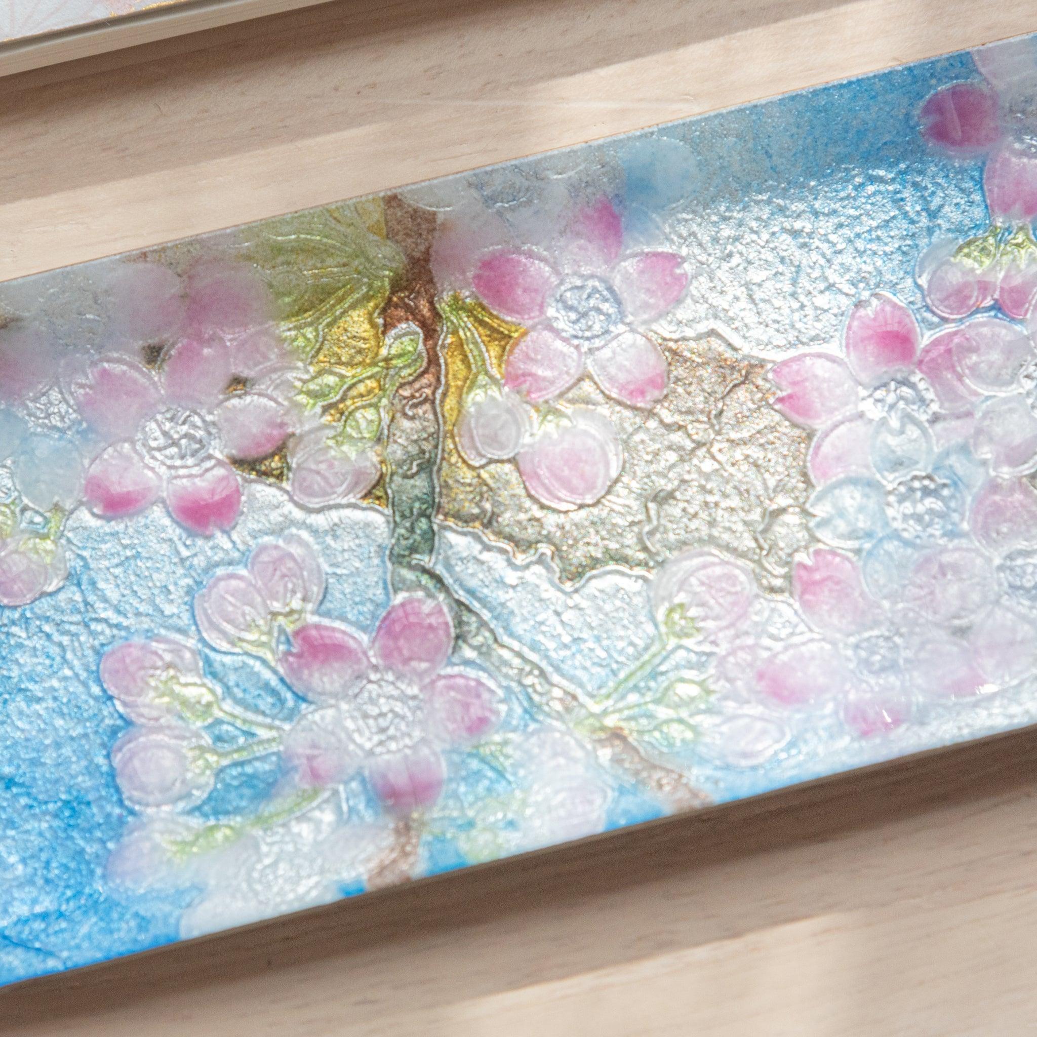 Rectangular tray with pink cherry blossom design, next to a fan-patterned notebook and blue glass pen on a light wooden surface.