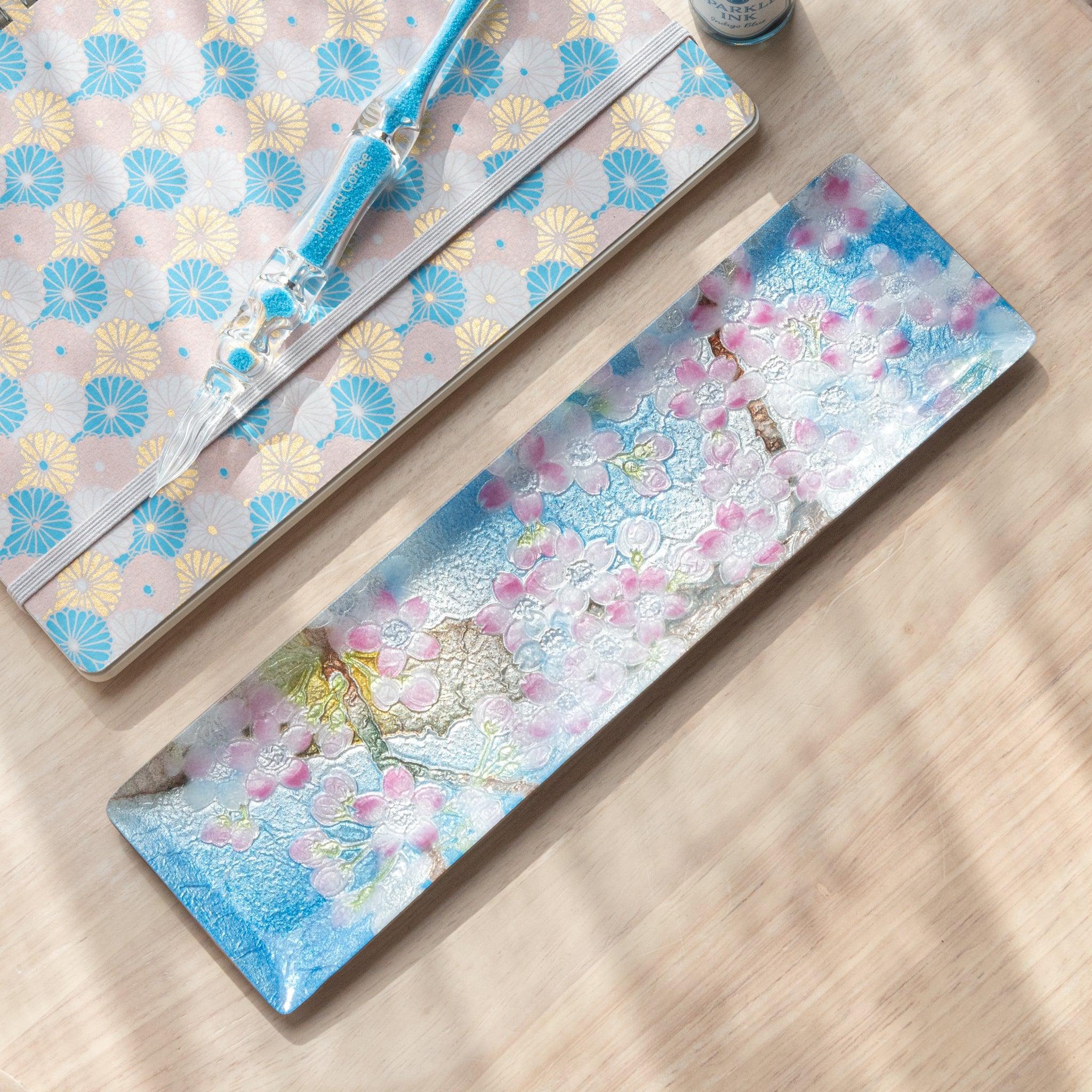 Close-up of cherry blossom pattern on a textured tray, featuring pink petals and branches with fine detailing on a blue background.
