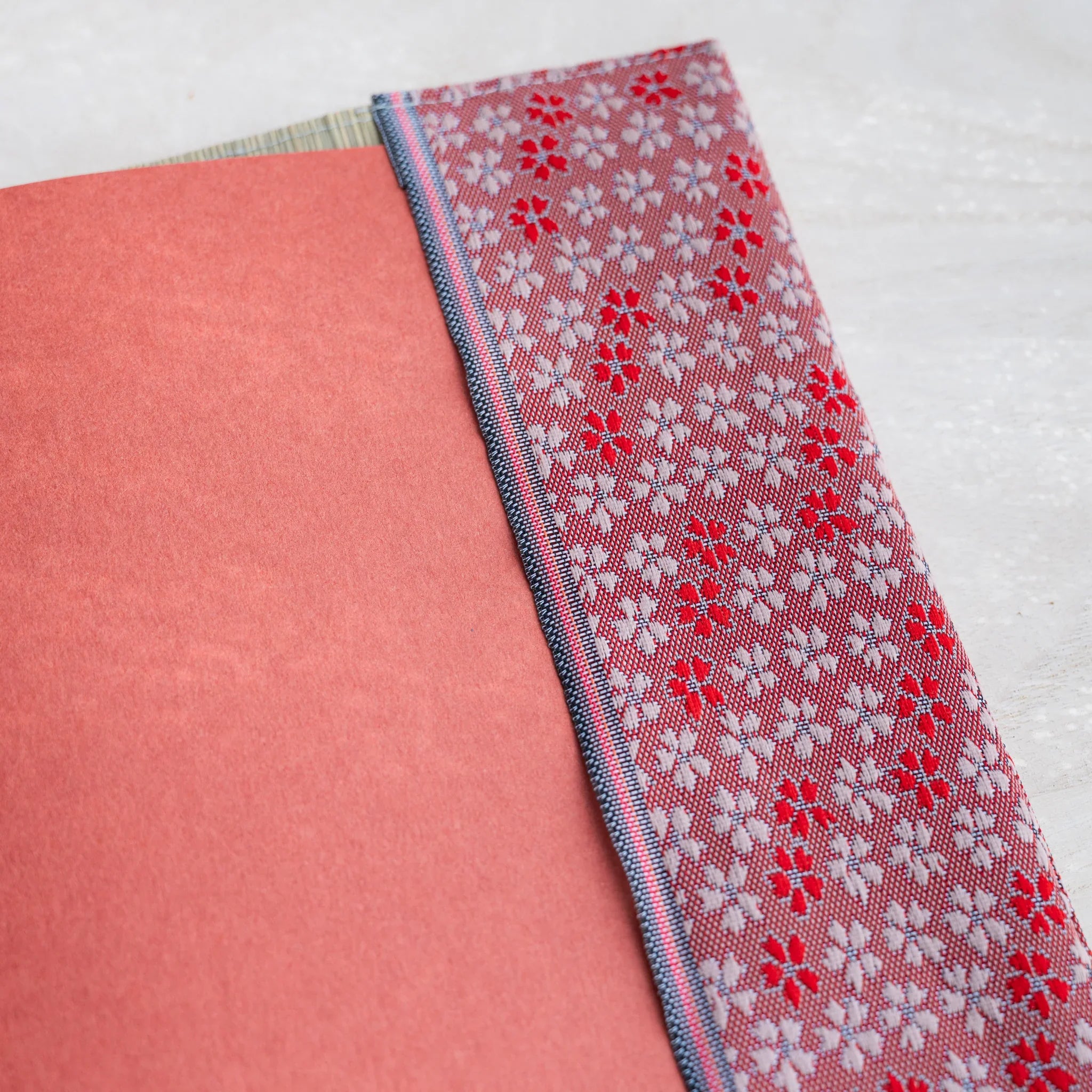 Close-up of pink and red cherry blossom brocade band beside a coral notebook cover inside the tatami book cover